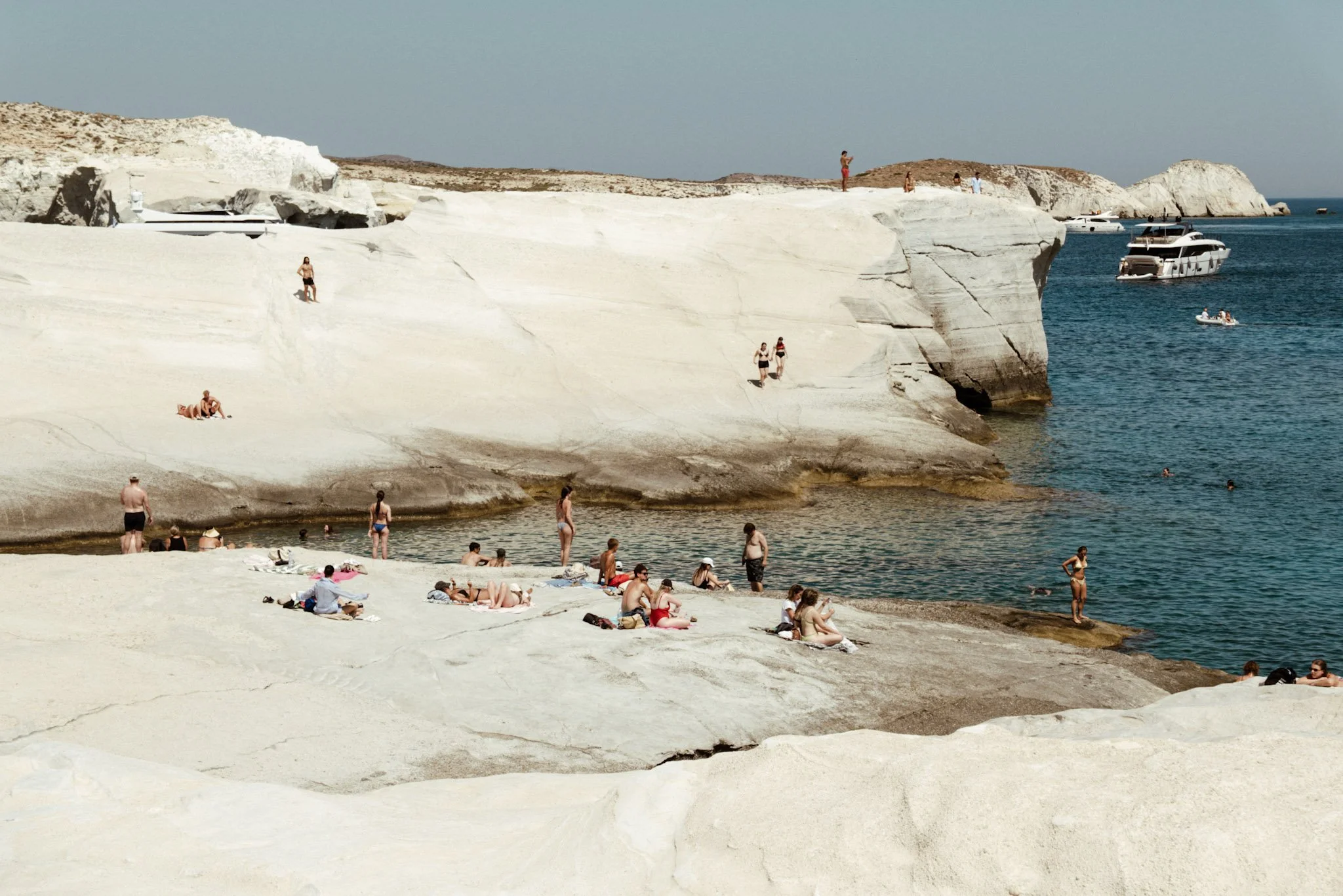 People relaxing and swimming at a beach with white cliffs and boats in the water.
