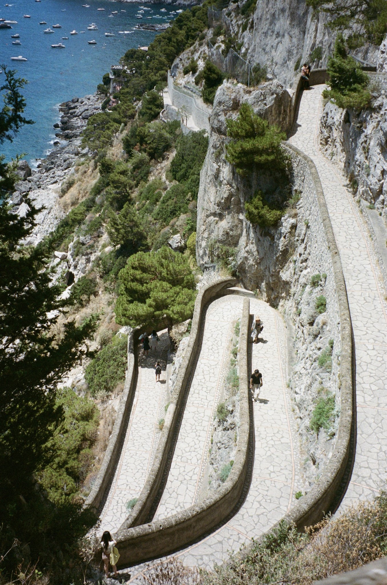 A coastal walkway with a narrow, winding path along a cliffside, overlooking a bay filled with boats. Several people are walking on the path, surrounded by greenery and rocky terrain.
