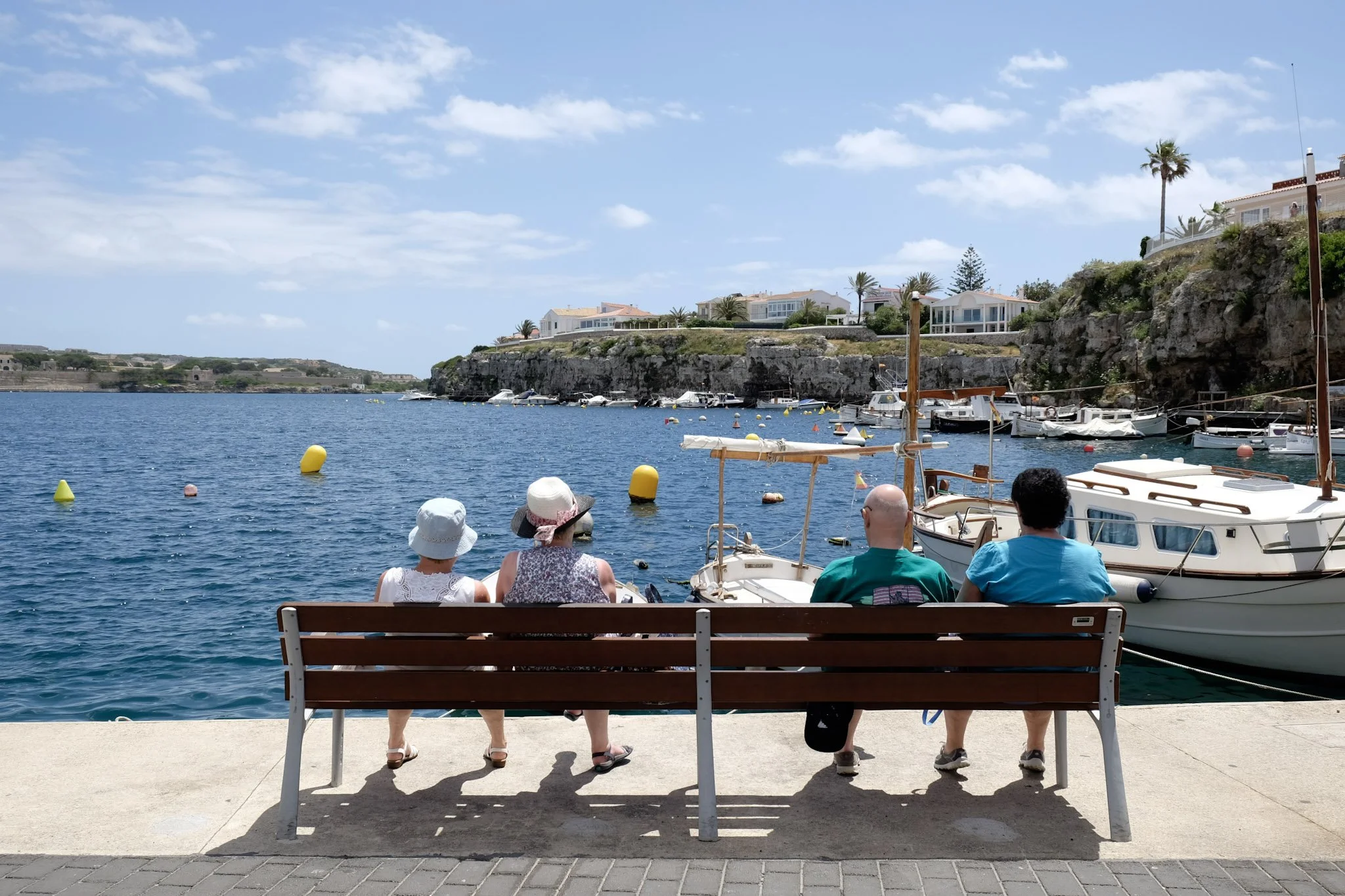 People sitting on a bench near a waterfront with boats docked, houses on a cliff, and a blue sky with clouds.