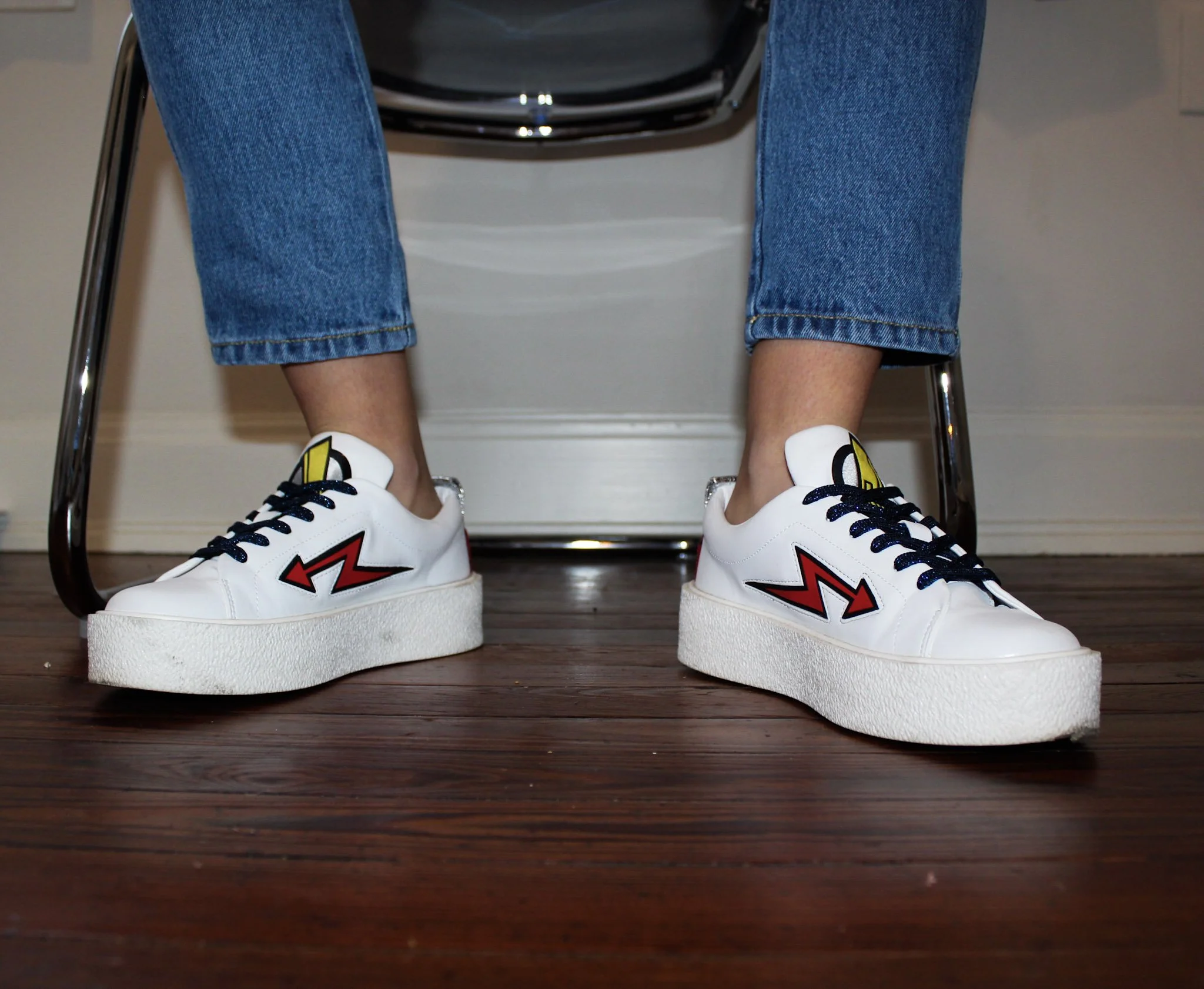 Close-up of a person's legs wearing blue jeans and sneakers, sitting on a chair with a black backpack visible in the background.