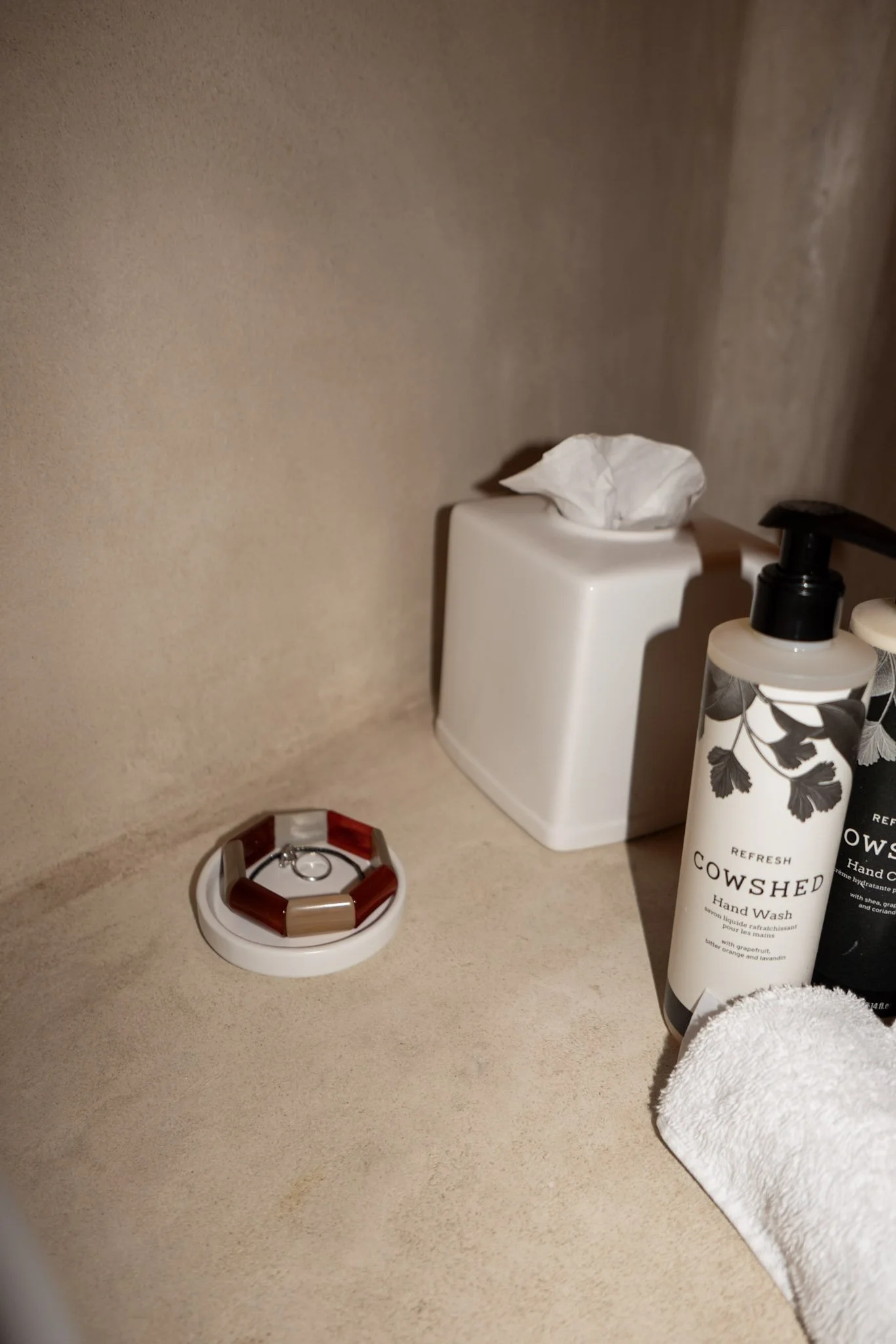 A beige countertop with a small white container with a tissue on top, a bottle of Cow Shed hand wash, a bracelet with brown and beige segments, a white towel, and part of a red and white item at the bottom left.
