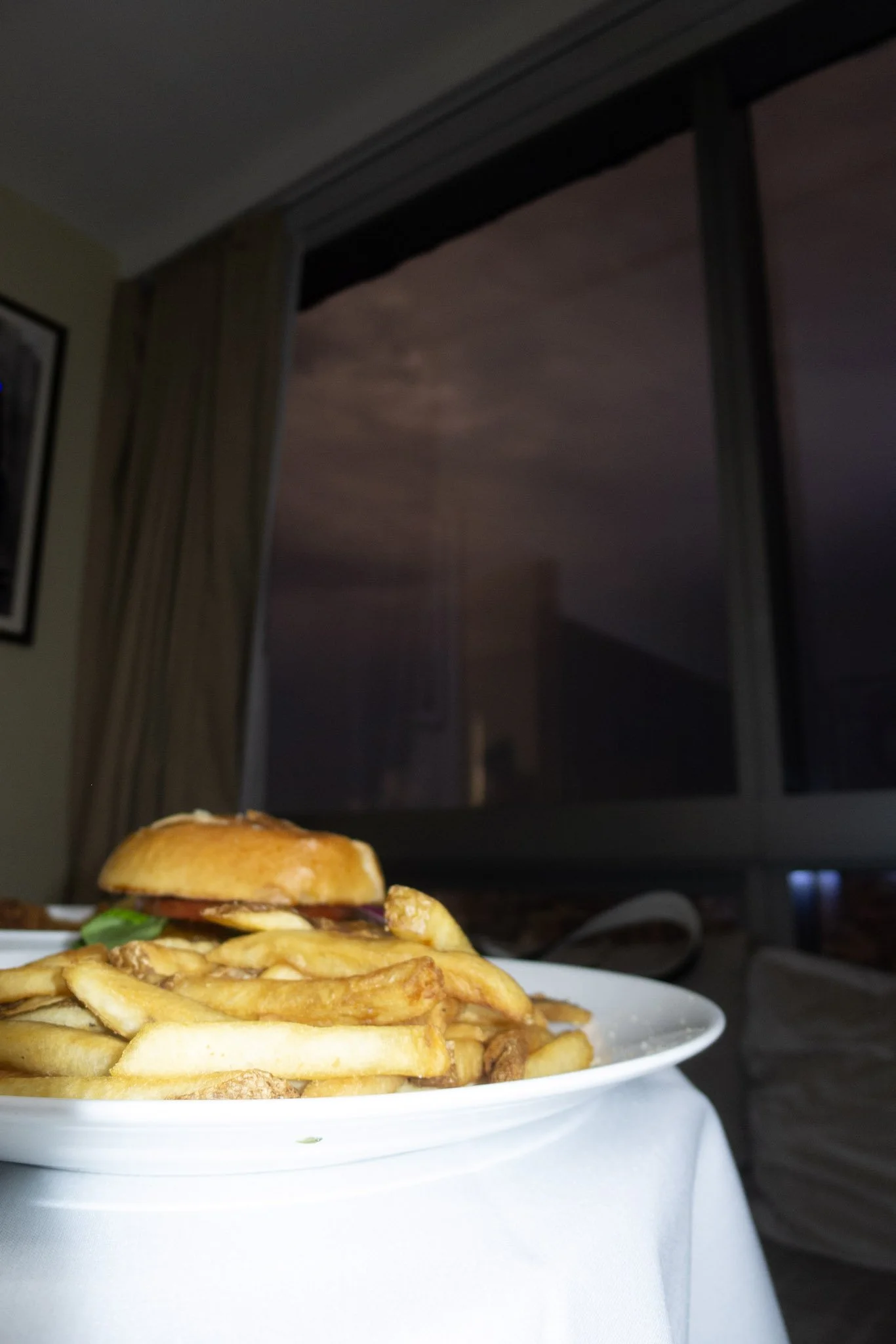 Close-up of a plate of French fries with a cheeseburger in the background, set on a white table near a window with curtains.