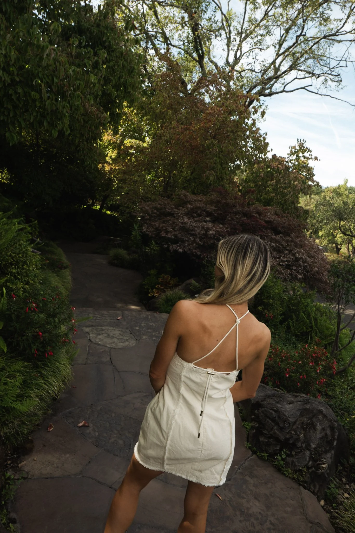 A woman in a white summer dress with spaghetti straps standing on a stone pathway in a lush garden surrounded by trees and plants, with her back turned and blonde hair.
