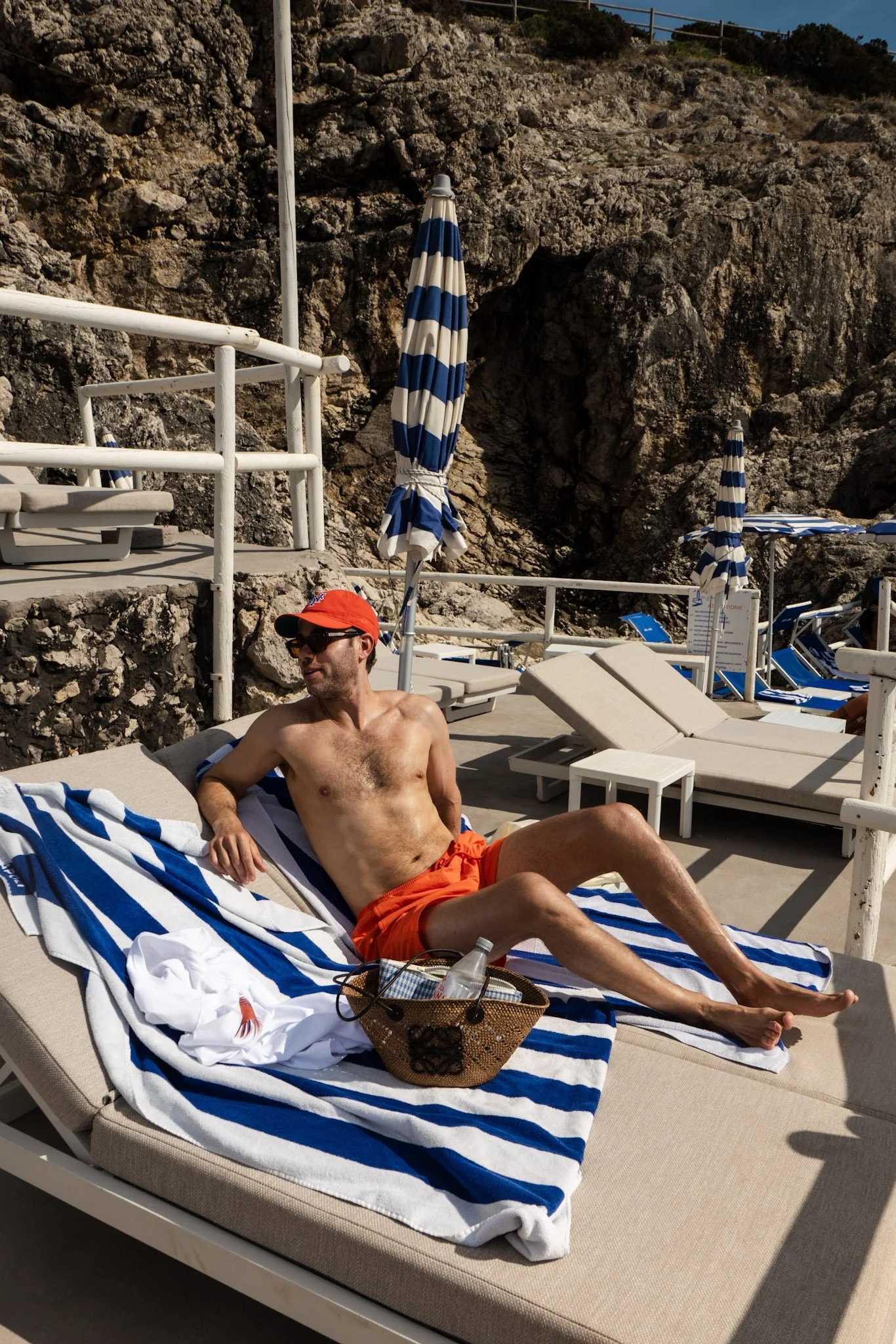 A man wearing orange swim trunks and a red cap is lounging on a striped towel on a sunbed at a beach. There are blue and white striped umbrellas and other sunbeds around him, with large rocky cliffs in the background.
