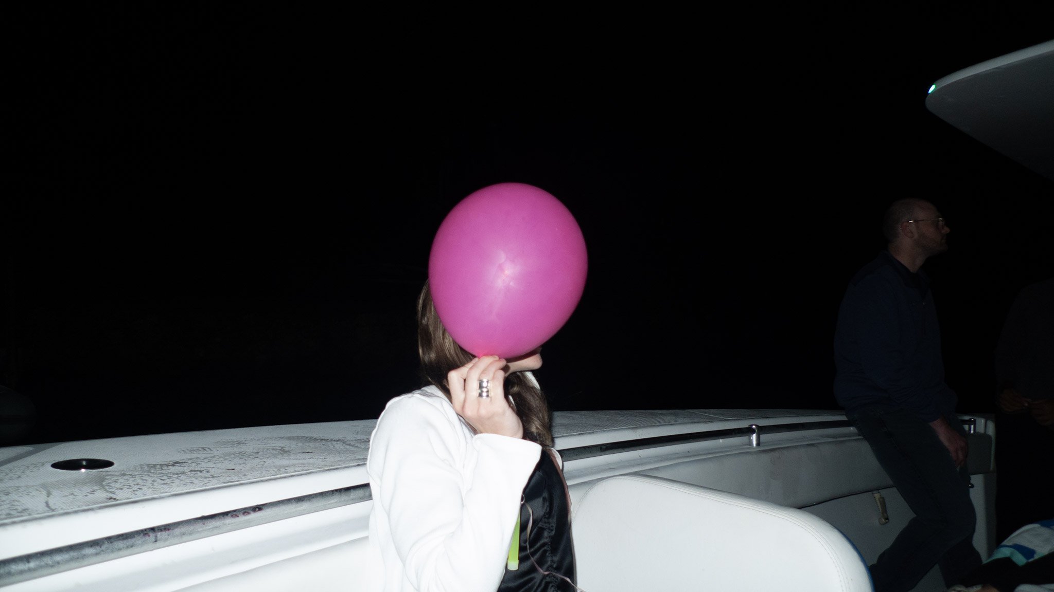 A person holding a pink balloon in front of their face on a boat at night, with another person standing nearby.