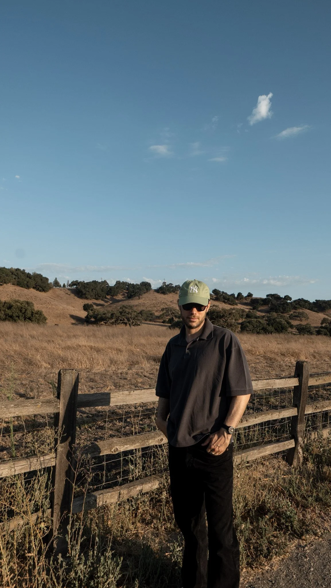 A man wearing a black jacket, black pants, a green baseball cap, sunglasses, and a watch stands outdoors near a wooden fence. Behind him are rolling hills with dry grass and scattered trees under a blue sky with a few clouds.