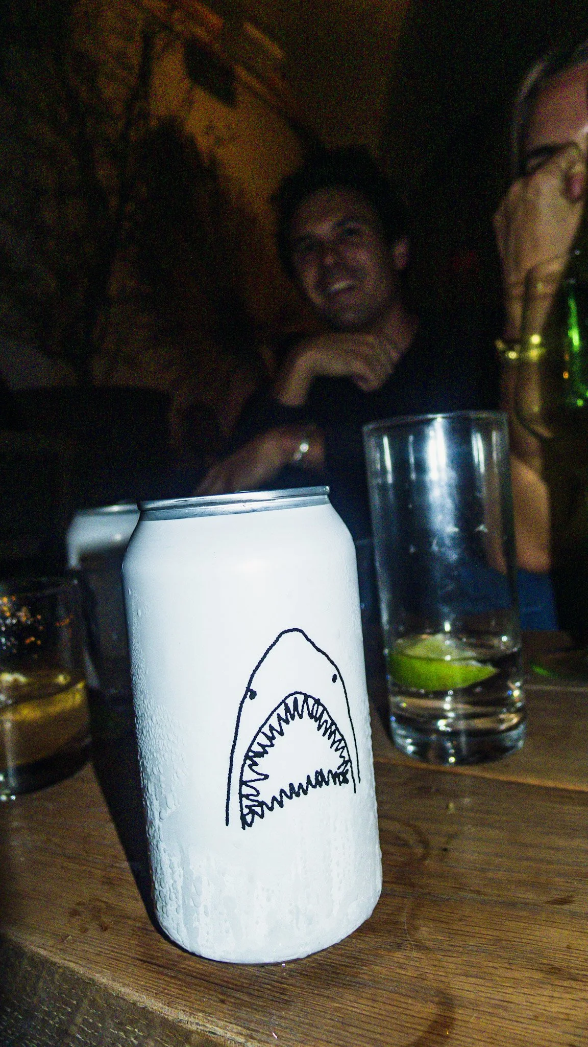 Close-up of a white can with a black design of a shark's open mouth, set on a wooden table along with glasses containing drinks, limes, and a person in the background at a dimly lit bar or restaurant.