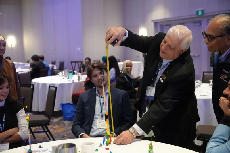 An elderly man in a suit demonstrating stacking colorful building blocks with a measuring tape at a conference or event, surrounded by seated adults and standing individuals in a banquet hall.