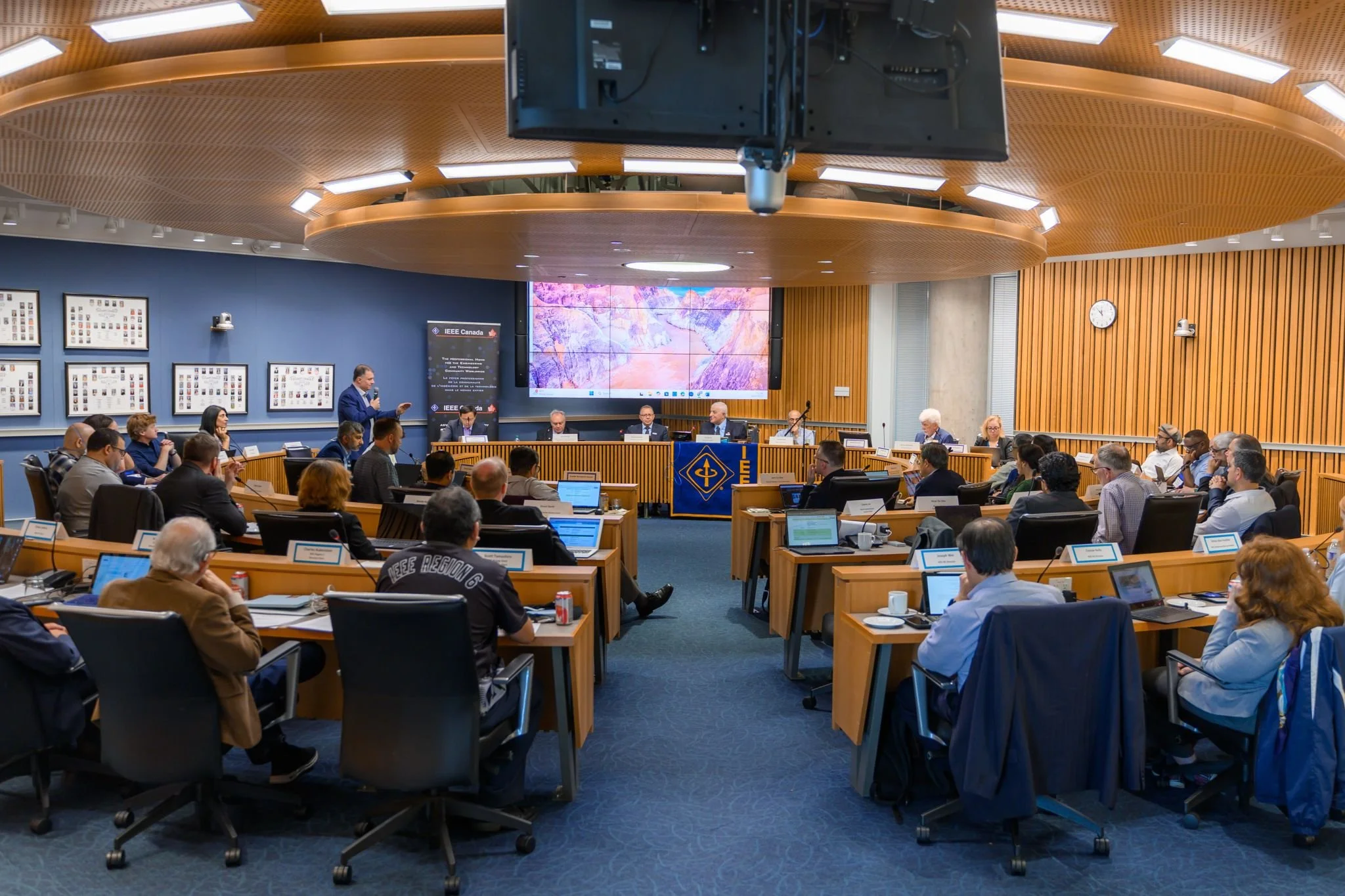 A large conference room filled with people seated at wooden tables, attending a meeting or presentation. There are six people sitting at a speaker's table at the front, with one person standing and speaking into a microphone. The room has wood-paneled walls, large display screens, and framed photographs on the walls, with a clock showing the time as 11:23.