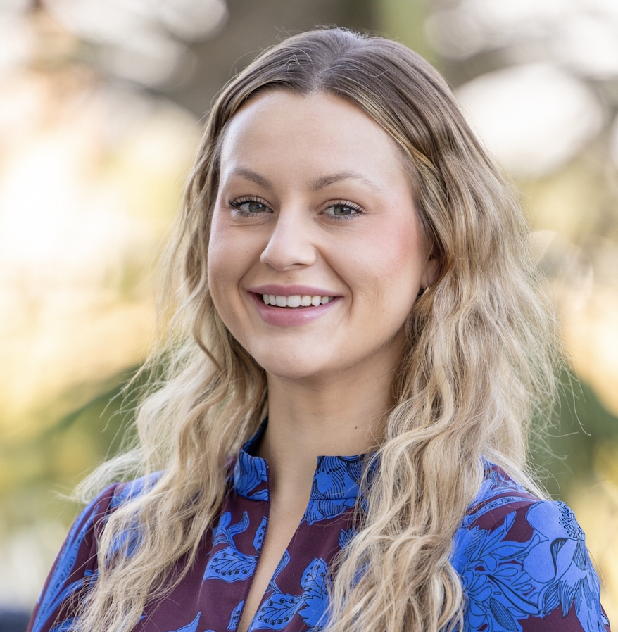 A young woman with long wavy blonde hair wearing a blue patterned blouse, smiling outdoors with blurred trees in the background.