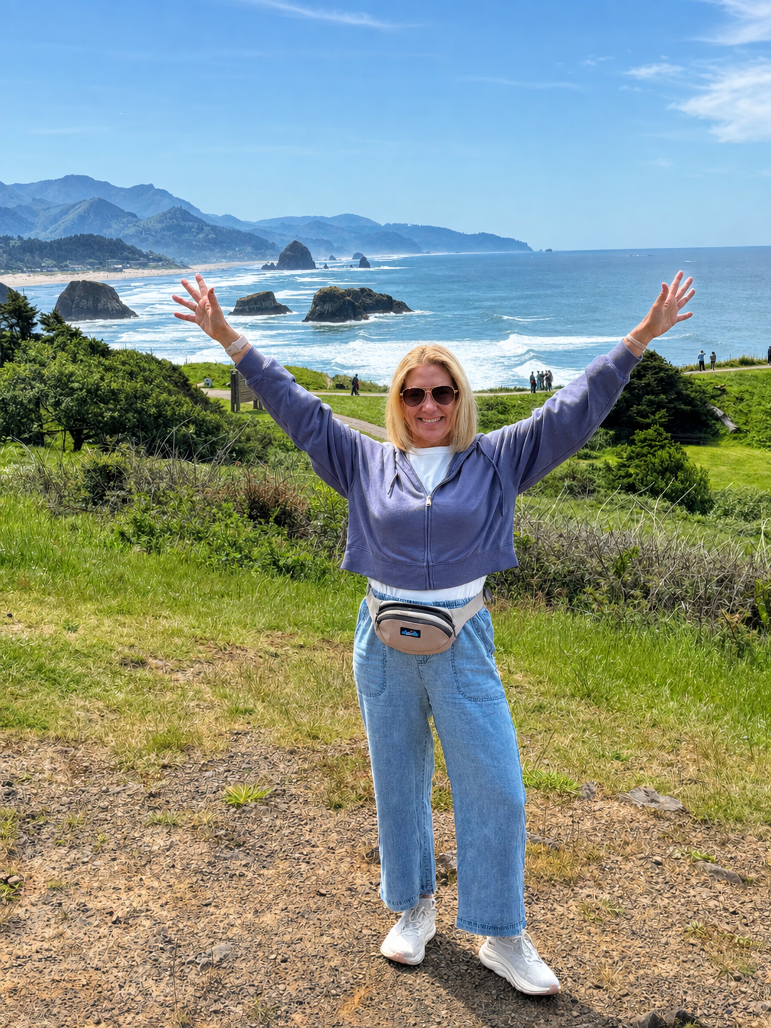 A woman standing outdoors on a grassy area with her arms raised, smiling at the camera, with a scenic ocean view and rocky coastline in the background.