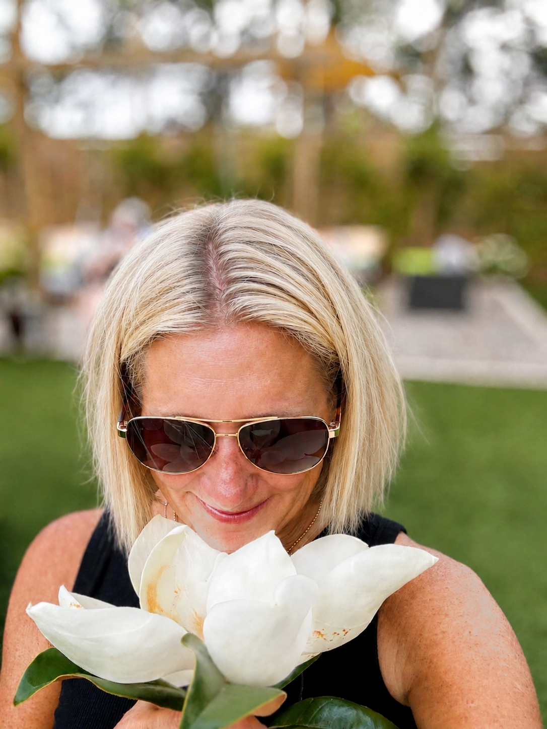 A woman with blonde hair wearing sunglasses holding a large white flower close to her face in an outdoor setting.