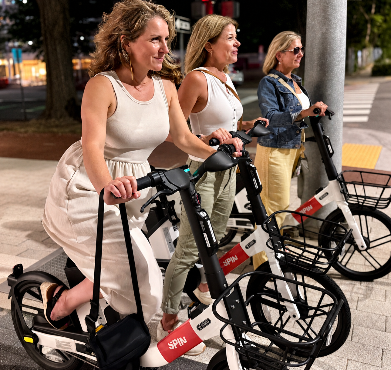 Three women wearing casual clothing, standing on electric bikes, waiting at a city crosswalk at night.