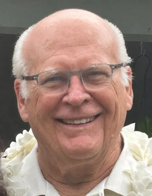 Close-up of an smiling elderly man with glasses, short white hair, and a floral necklace.