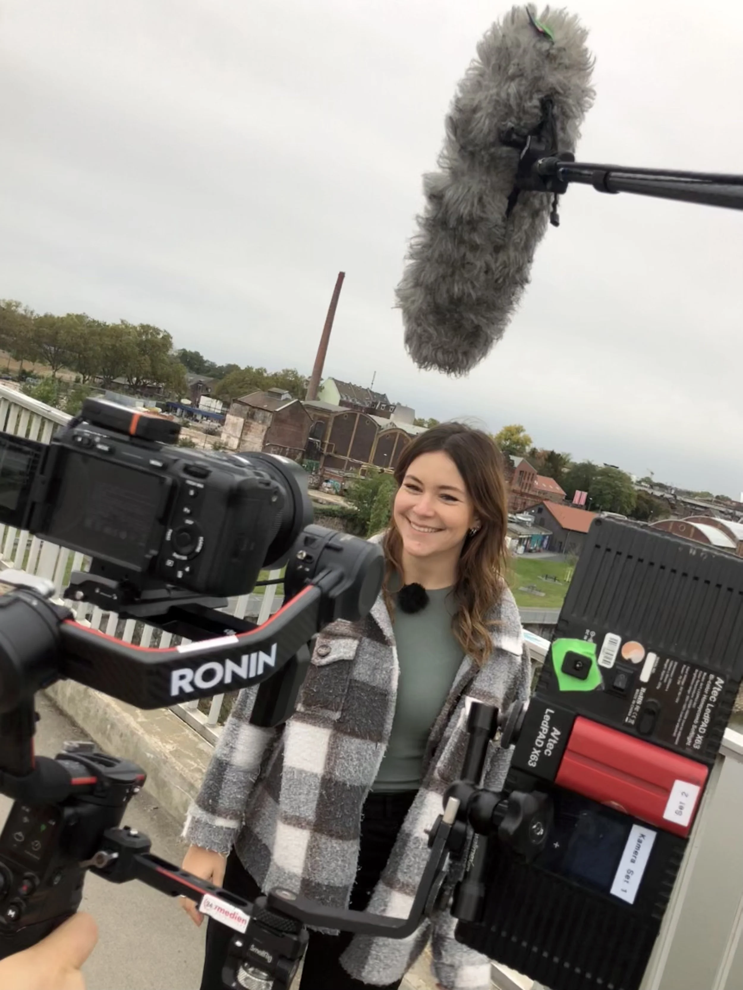 Eine lachende junge Frau steht vor einer Kamera, während ein großer Mischsound-Mikrofon über ihr schwebt. Es scheint, als würde sie bei einem Videodreh oder Interview mit dem Hintergrund von Häusern und Bäumen im Freien sein.
