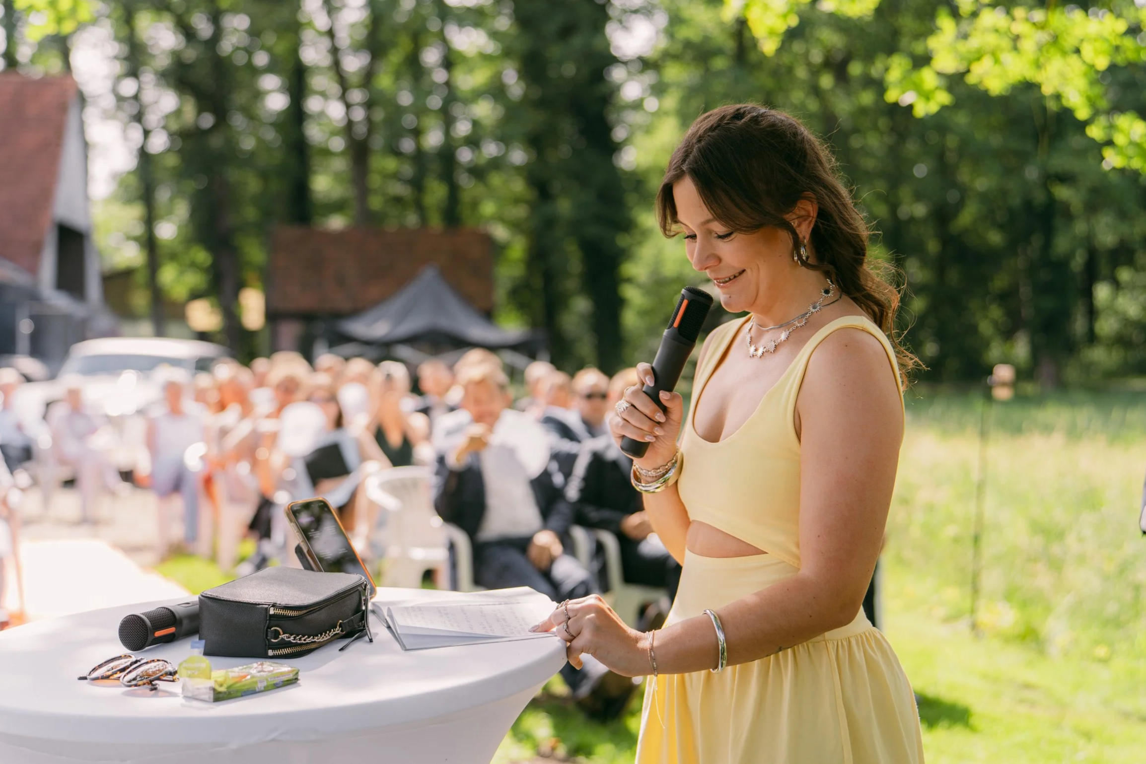 Frau in gelbem Kleid hält Mikrofon beim Reden bei einem Outdoor-Event, im Hintergrund sitzt eine Gruppe von Menschen, die zuhörn. Tische mit Gegenständen, unterwegs in einem grünen Park.