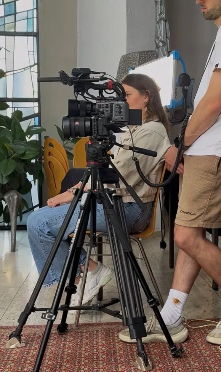 Eine Person sitzt vor einer Kamera auf einem Stativ, während eine zweite Person dahinter steht und eine Kabelverbindung hält. Es scheint eine Aufnahmesituation in einem Raum mit Pflanzen und Fenstern im Hintergrund zu sein. Filme werden produziert