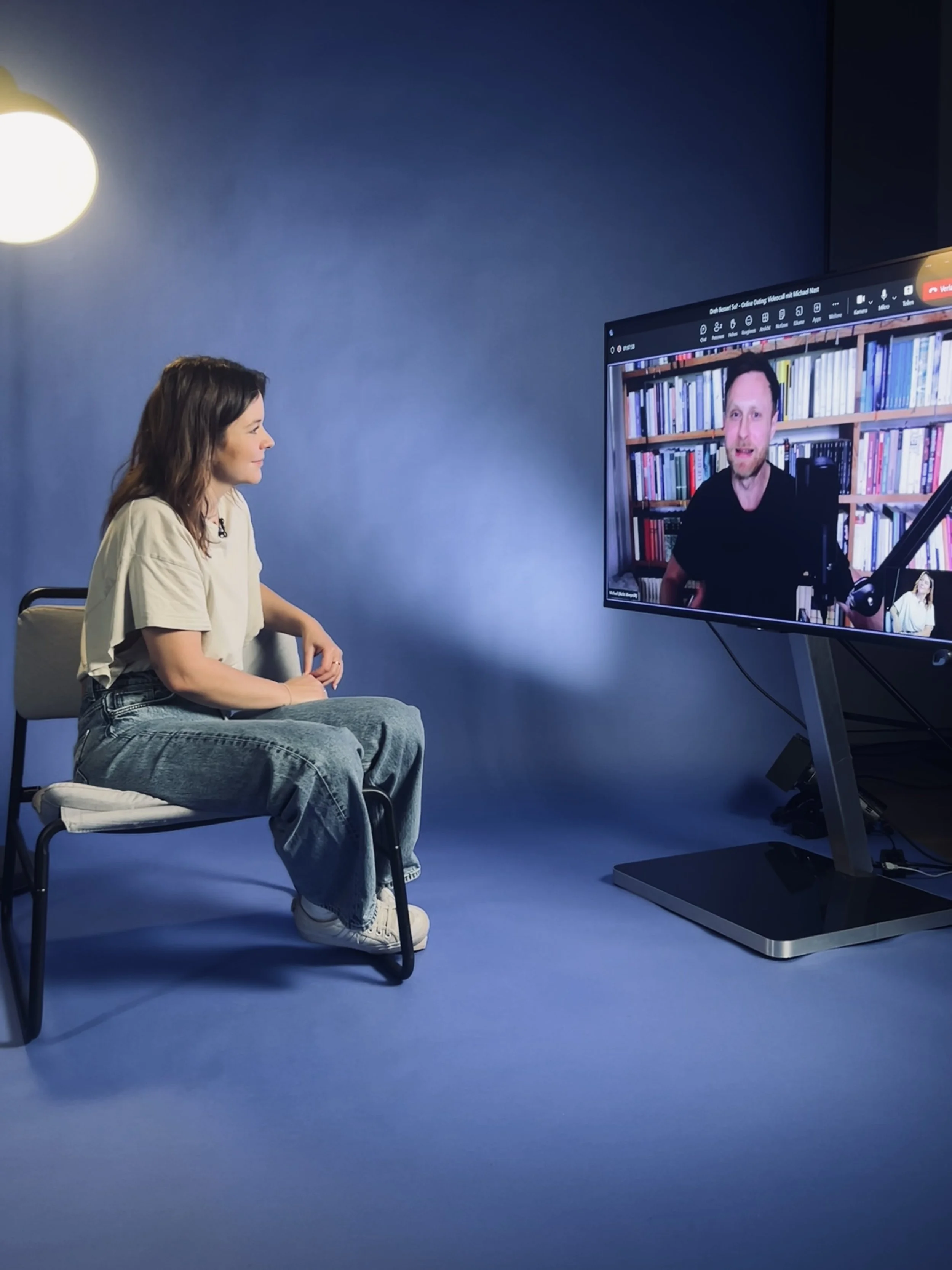 Eine junge Frau sitzt auf einem Stuhl vor einem blauen Hintergrund und schaut auf einen Bildschirm, auf dem ein Mann in einer Bibliothek spricht.