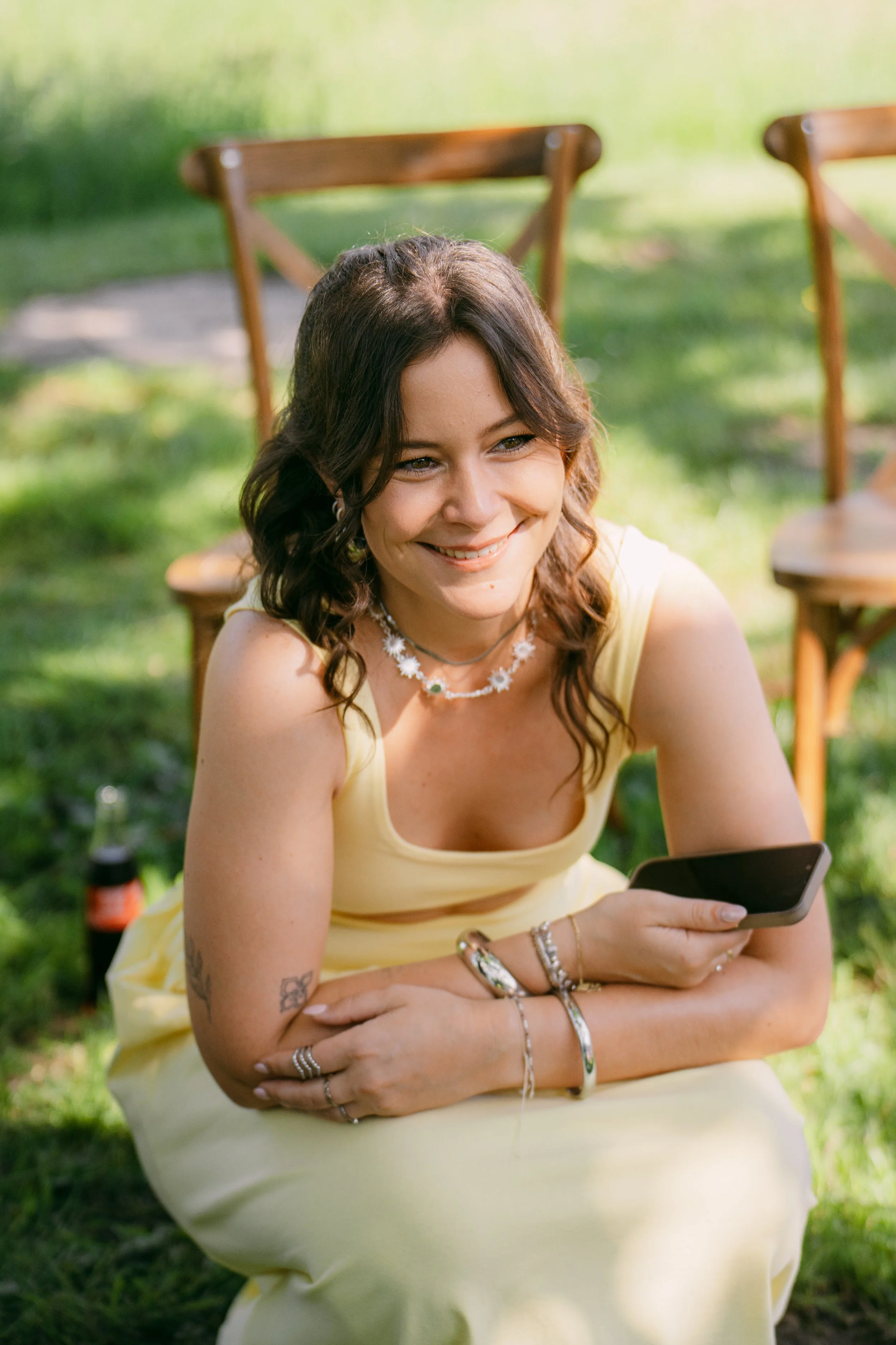 Junge Frau mit dunklen, gewellten Haaren, trägt ein gelbes Kleid und Schmuck, sitzt im Garten und schaut lachend auf ihr Smartphone.