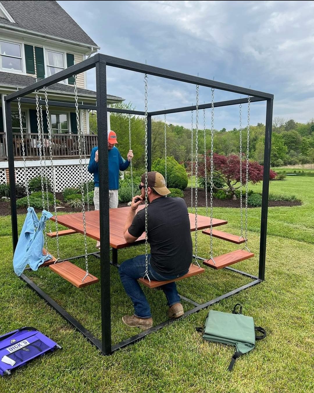 Two men are assembling a large outdoor lounge swing with a black metal frame and wooden seats in a backyard with green grass, trees, and a house with a deck. One man is sitting on the swing talking on the phone, while the other man stands nearby adjusting the frame. Tools and a folded cloth are on the ground.