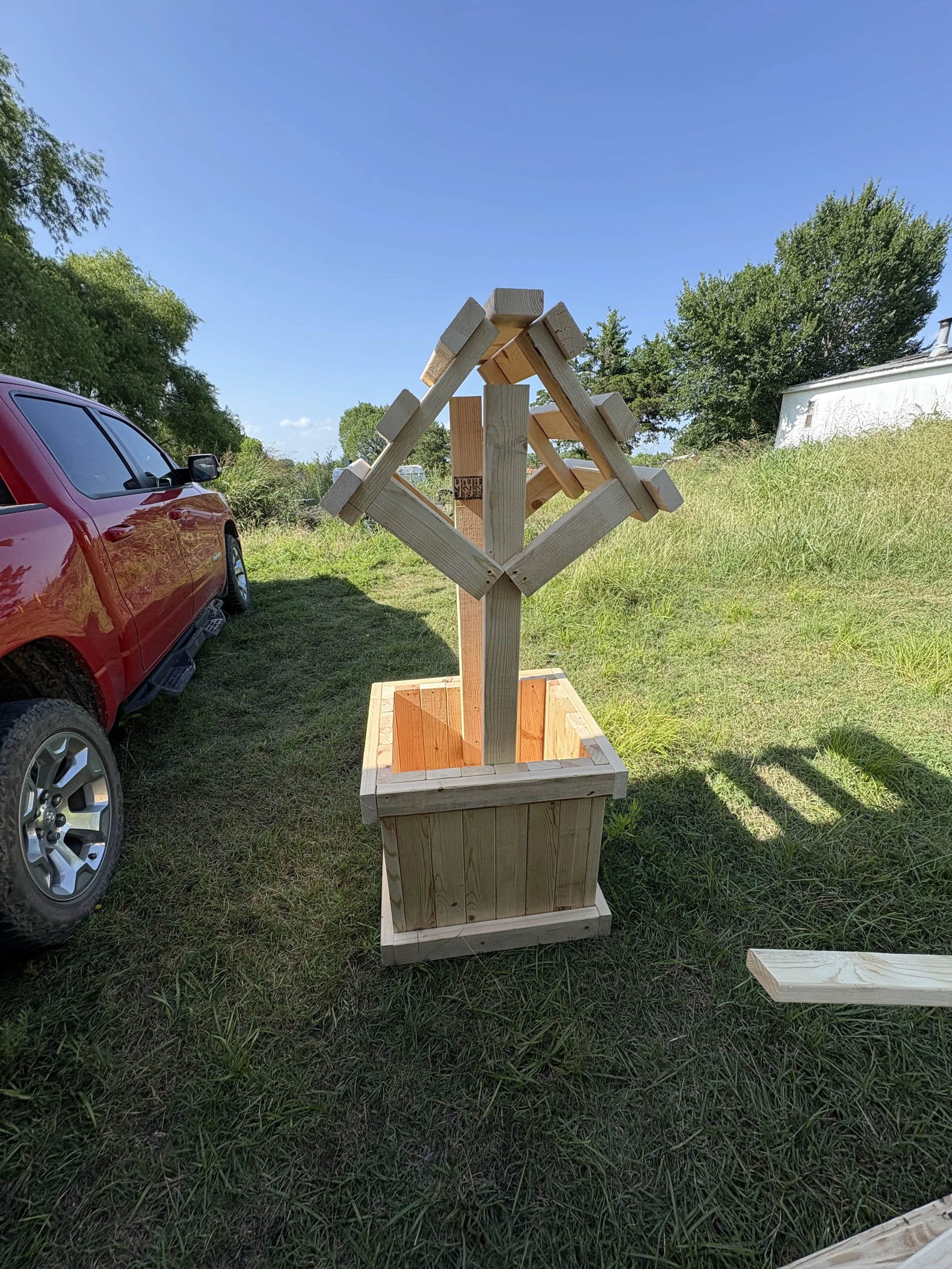 A wooden birdhouse on a stand in a grassy outdoor area with a red pickup truck to the left and trees in the background.