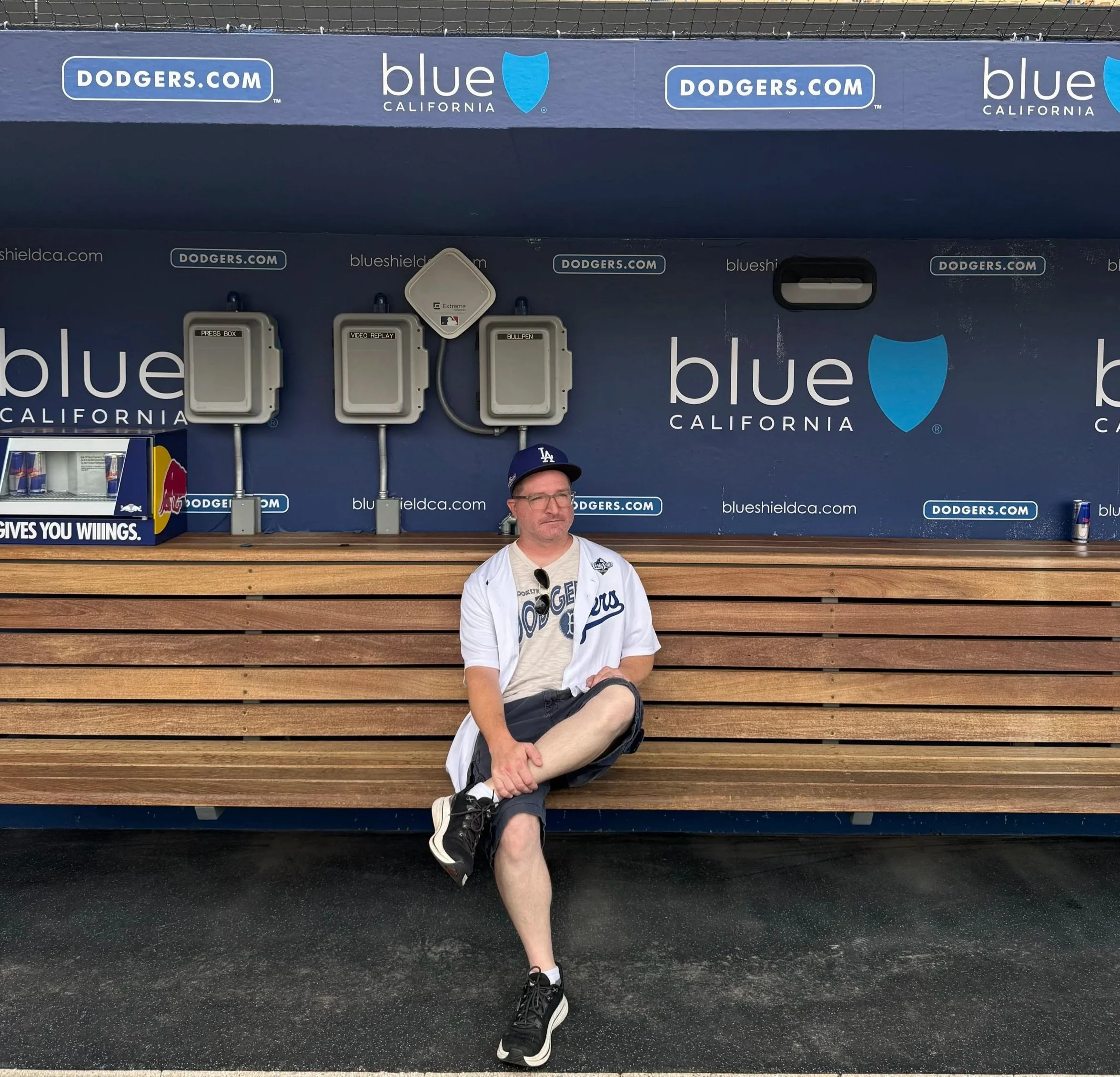 A man sitting on a wooden bench at a baseball stadium, wearing a Los Angeles Dodgers t-shirt, baseball cap, and sunglasses. Behind him is a blue wall with advertisements for Dodgers.com and Blue California, with electronic equipment mounted above.