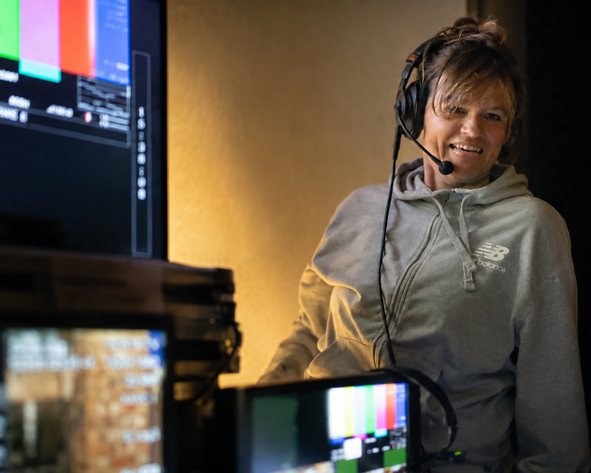 A man with long hair and a gray hoodie wearing a headset smiling while working in a studio with monitors and equipment.