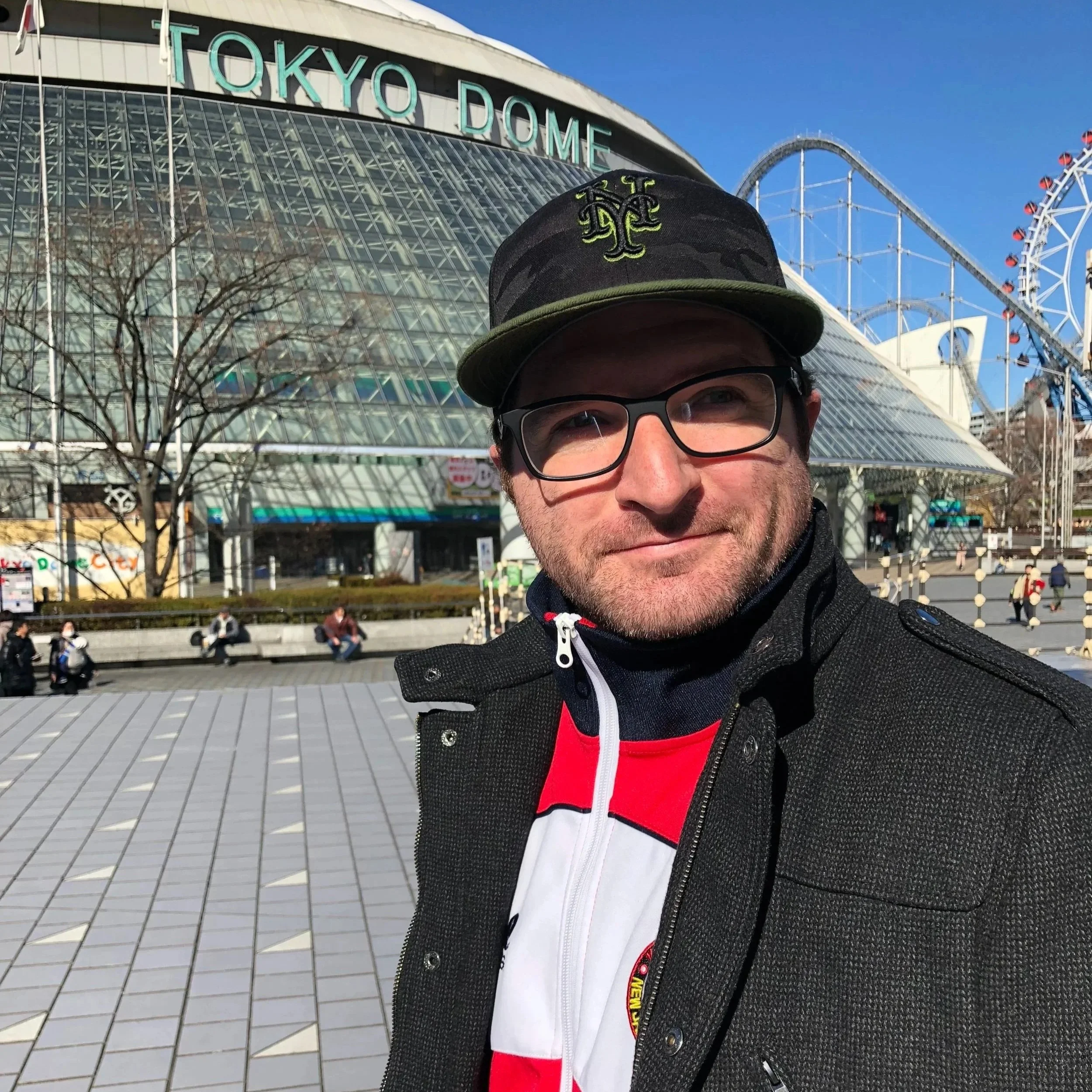 A person wearing glasses, a black hat with green embroidery, and a black and red jacket standing in front of Tokyo Dome and a roller coaster with a clear blue sky.