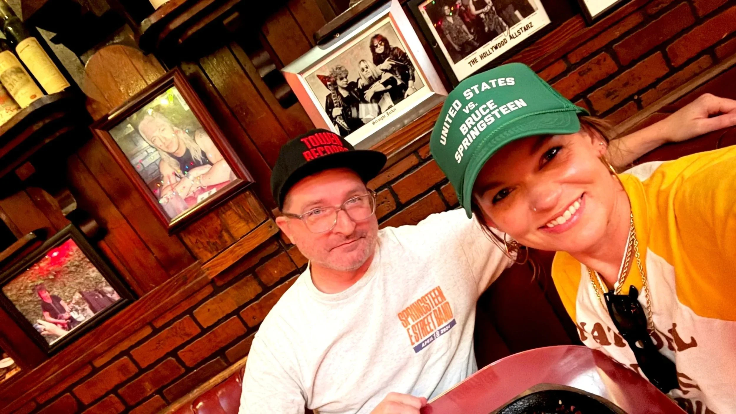 Two people, a man with glasses and a woman with a green cap, taking a selfie inside a restaurant with brick walls and framed pictures in the background.