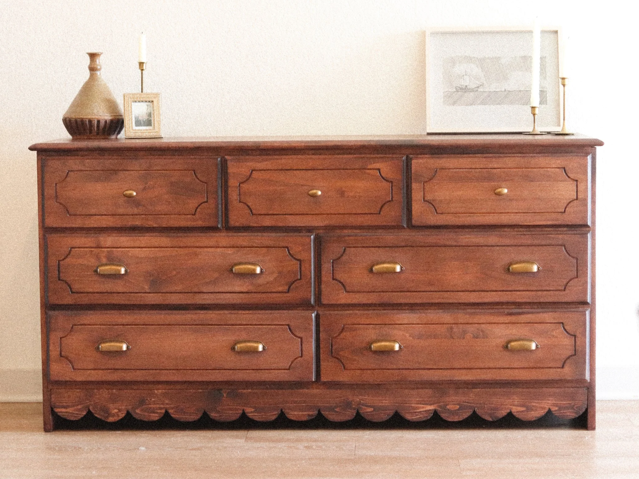 A vintage wooden dresser with six drawers, brass handles, and a scalloped bottom edge, situated against a beige wall.