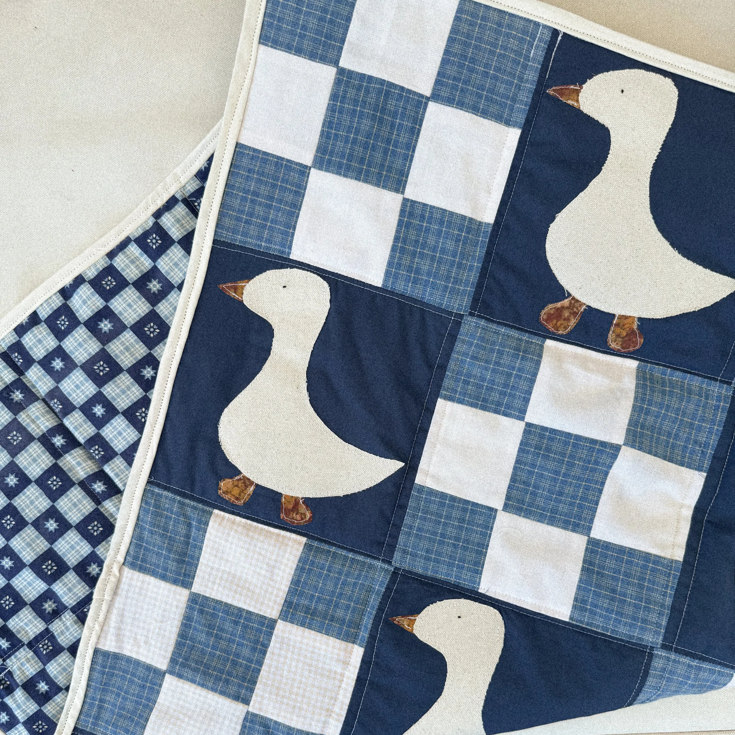 Close-up of a quilt with a penguin design, made of various blue and white checkered and plaid fabric patches, on a neutral background.