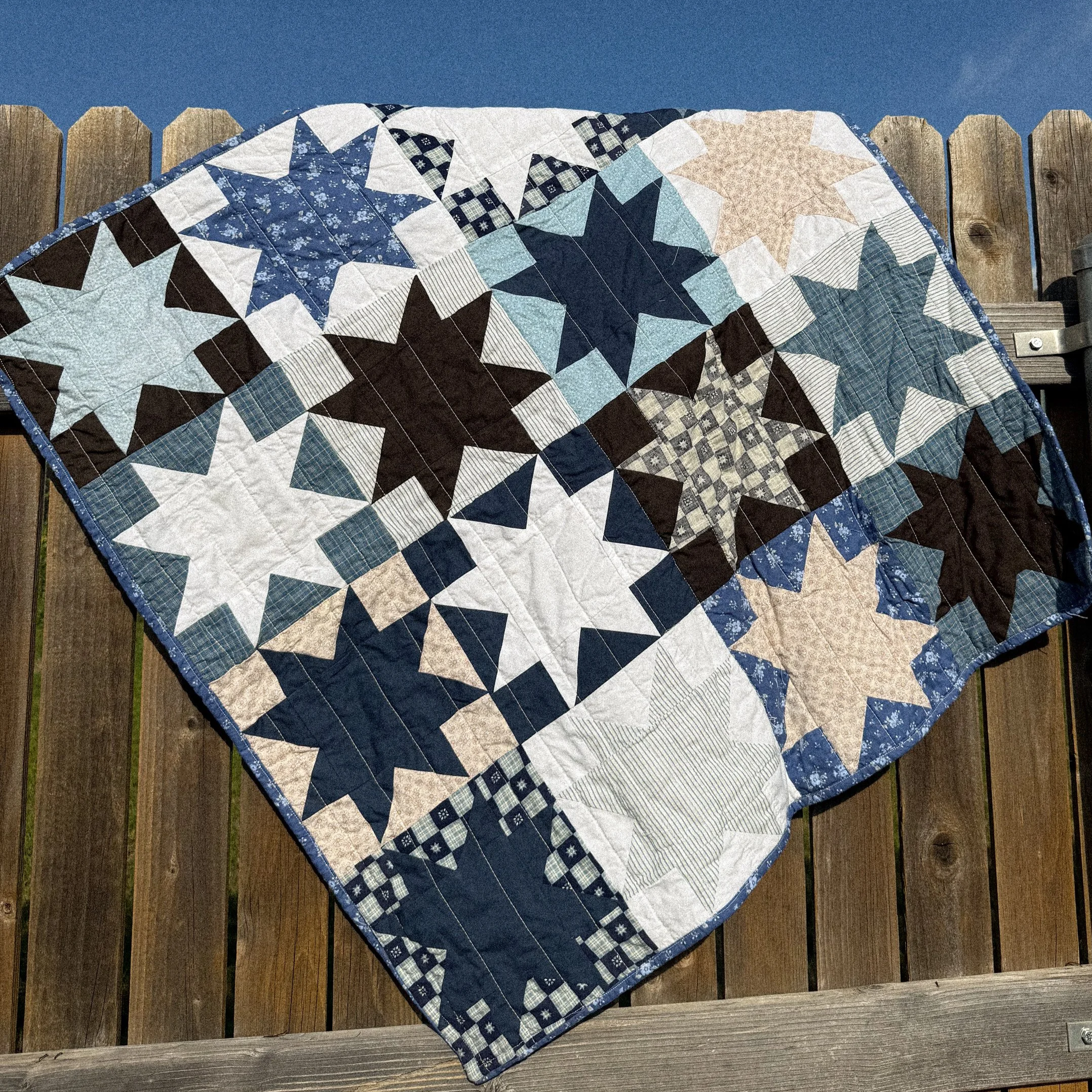 A quilt with a blue, white, and black star pattern draped over a wooden fence against a blue sky.