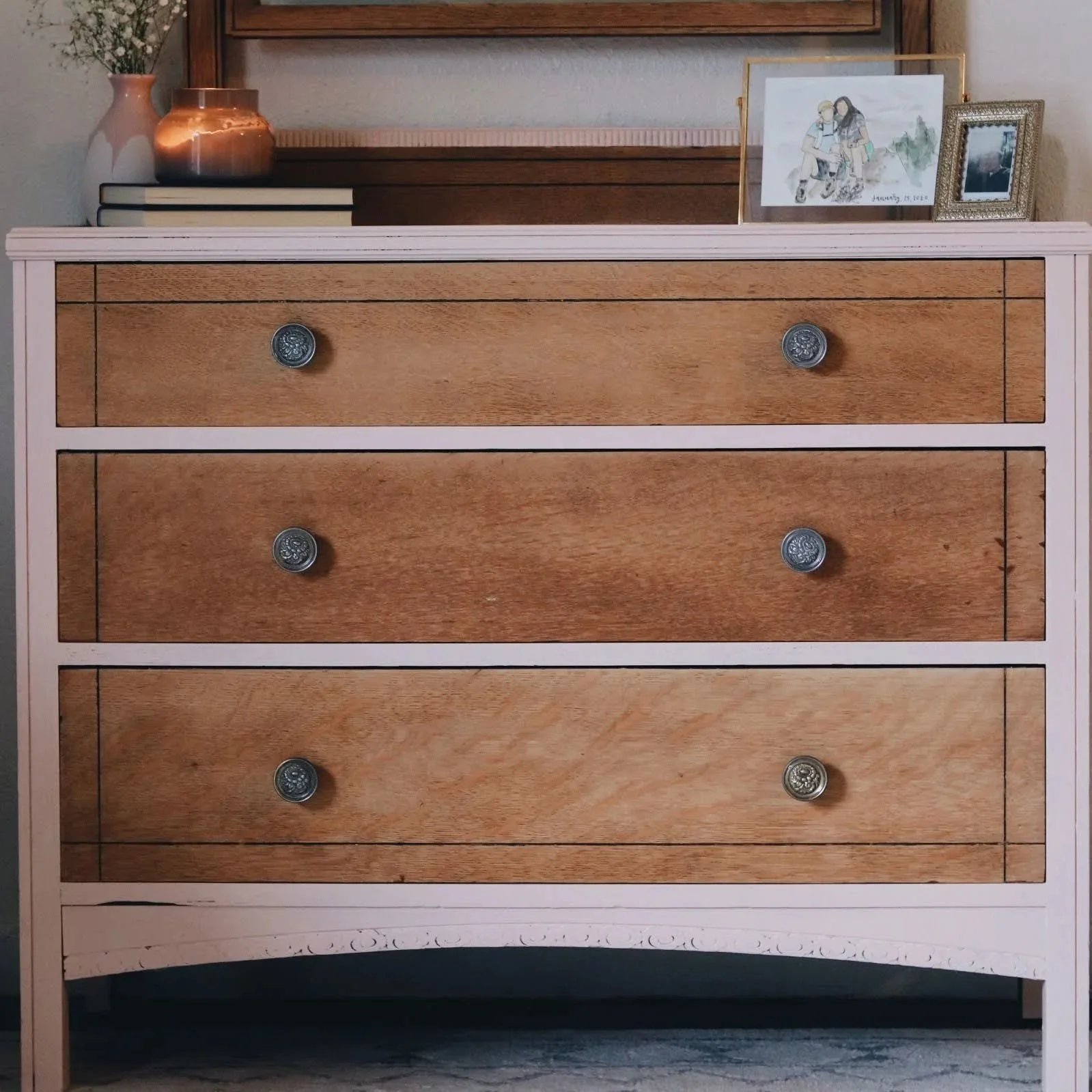 A wooden dresser with multiple drawers, a pink and white painted frame, decorated with a vase of flowers, framed artwork, and small decorative items on top.