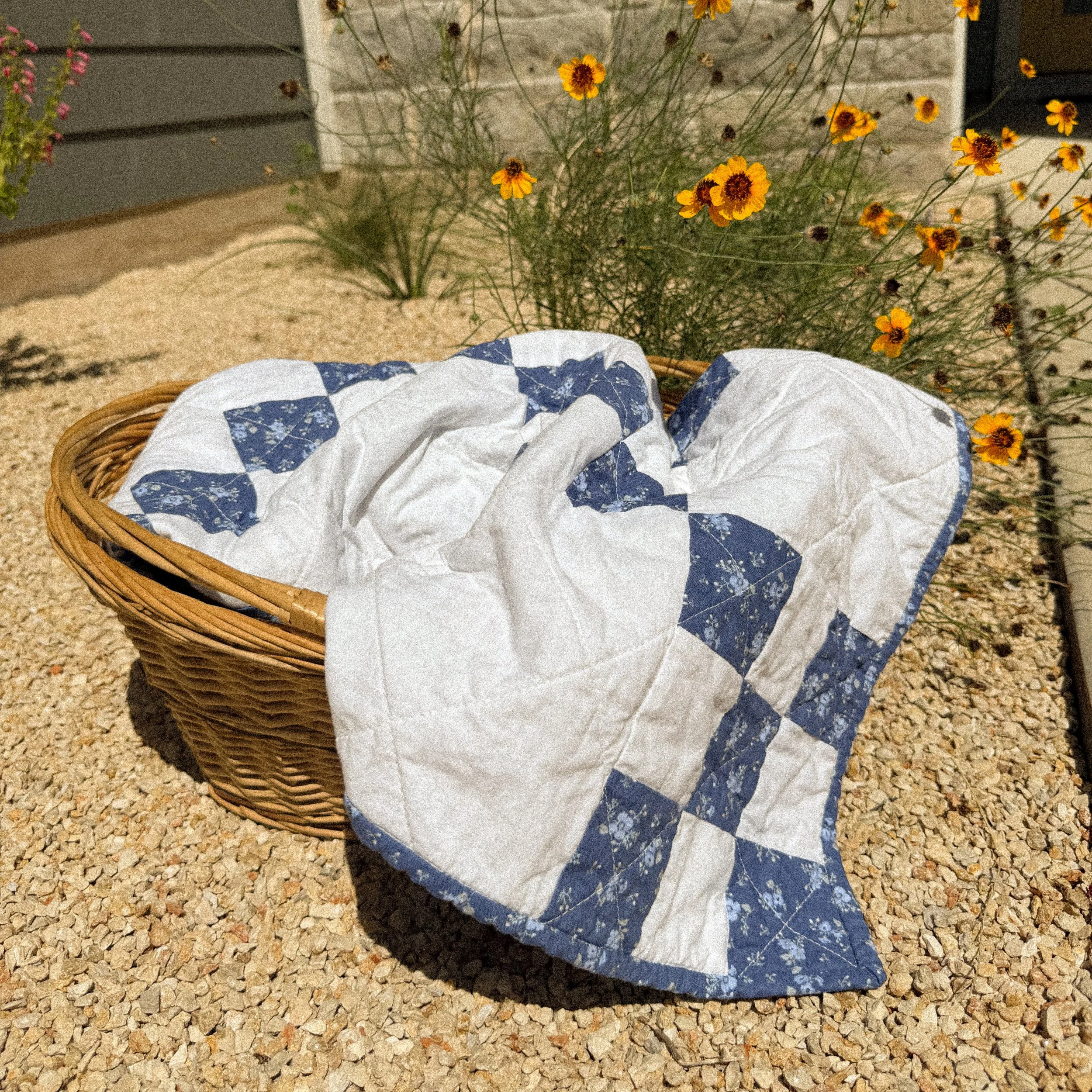 A wicker basket lined with white cloth with blue patterns, placed on a gravel surface with yellow flowers and plants in the background.