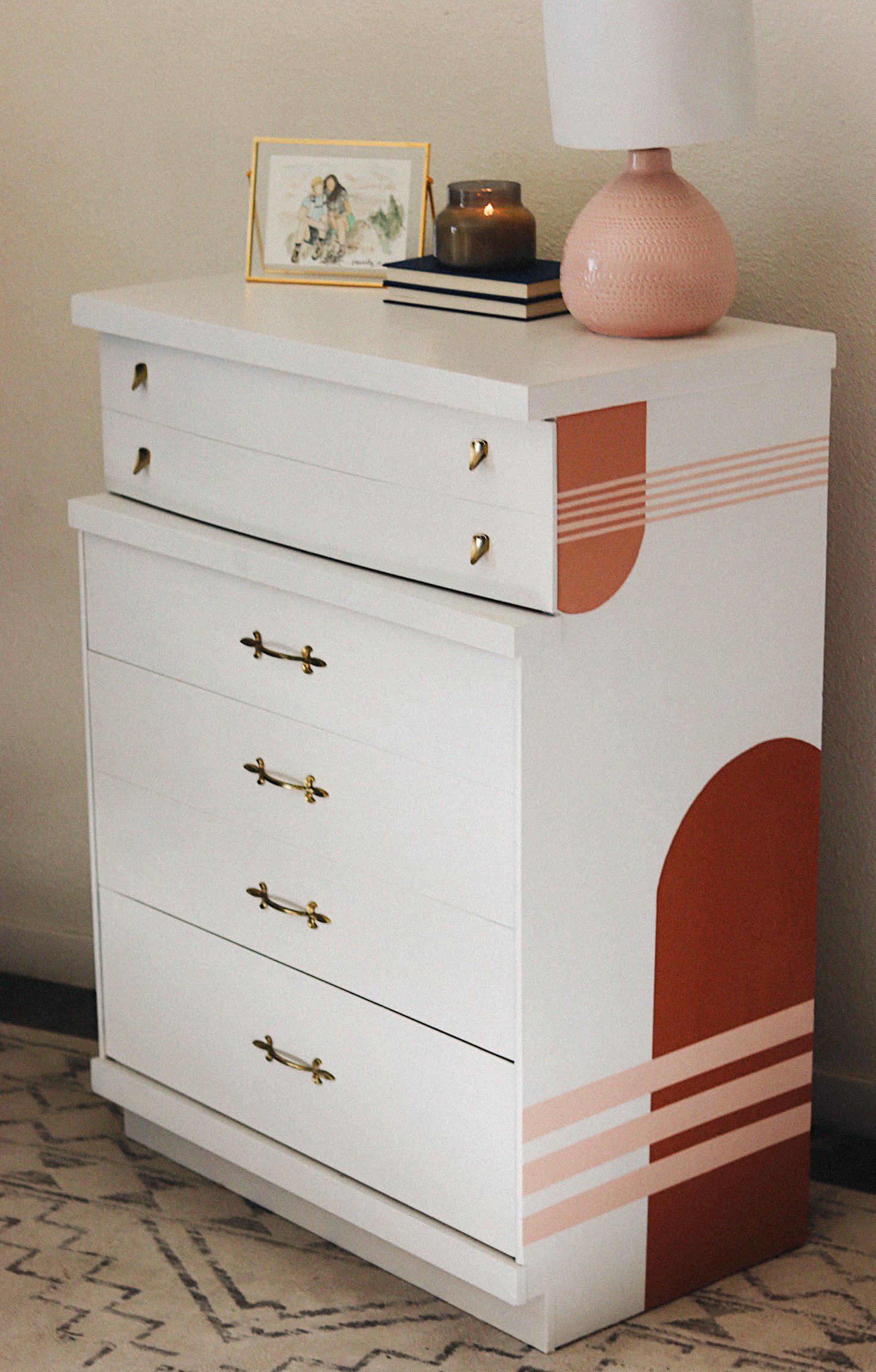 White dresser with gold handles, pink and brown geometric and striped design on the side, on a patterned rug.