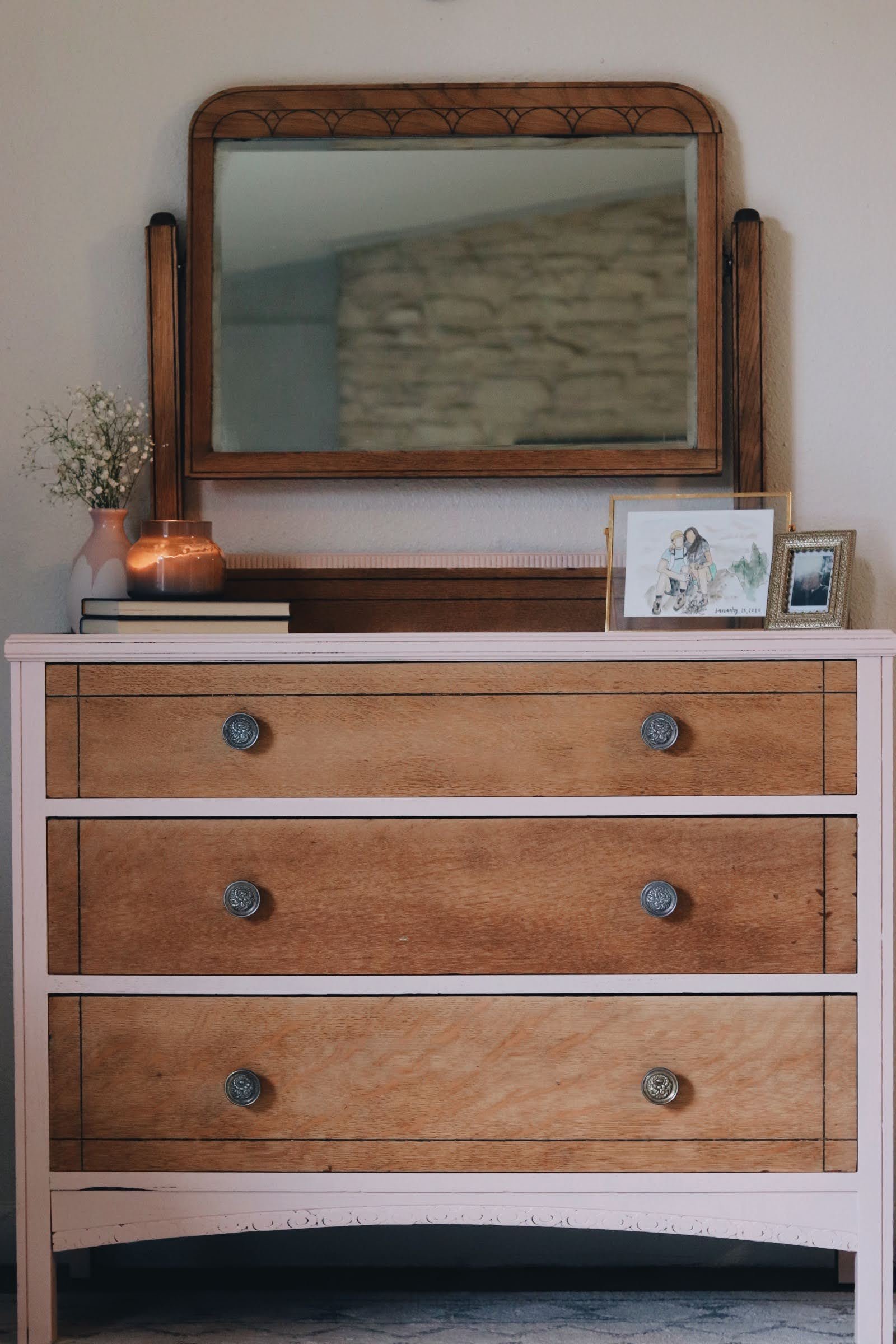 A wooden dresser with five drawers and silver knobs, topped with a mirror, a framed drawing of two people, a framed photograph, a one white and one wooden vase, and a small copper container.