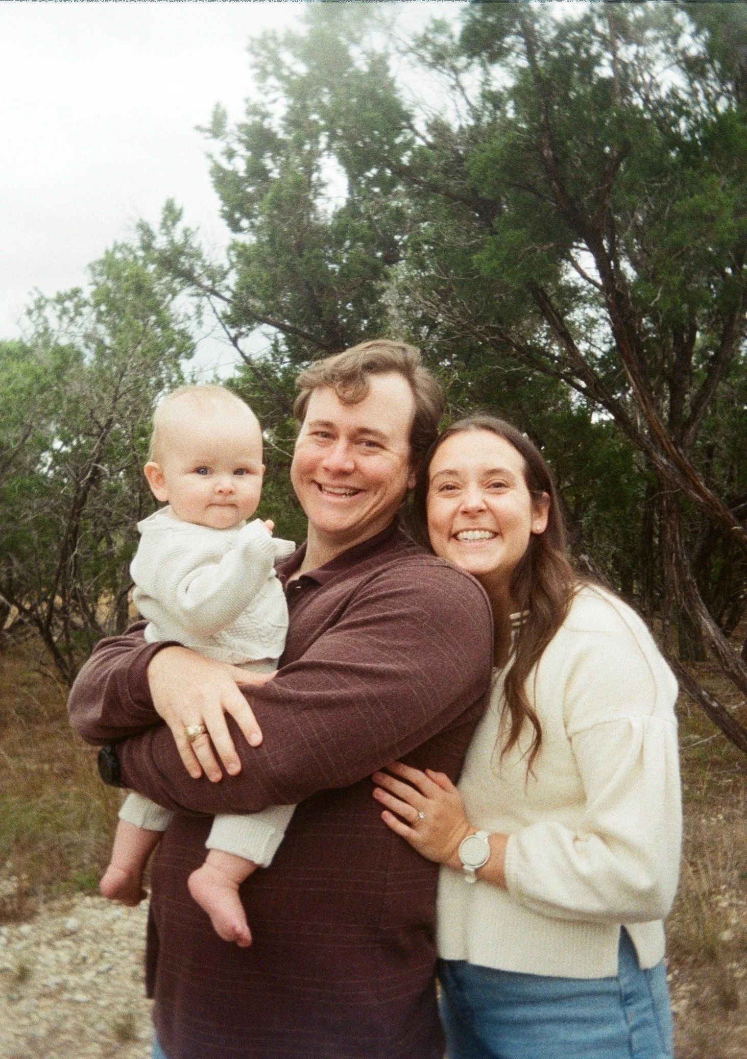 Family of three outdoors, a woman and man smiling, woman wearing light-colored sweater, man holding a baby girl, trees in the background.