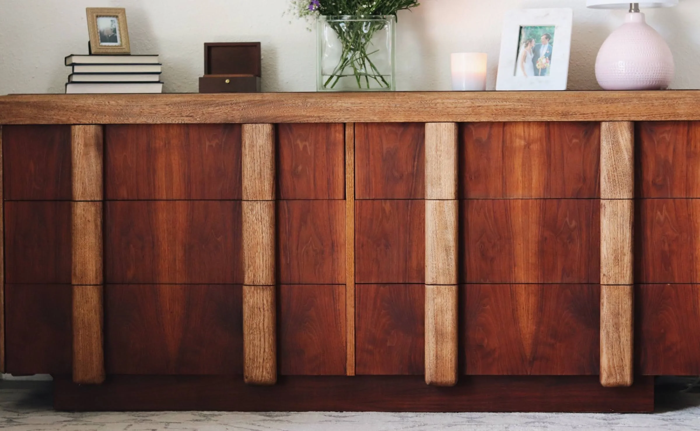 A wooden cabinet with multiple drawers, topped with various decorative items including a stack of books, a picture frame, a small box, a glass vase with purple flowers, a framed photo of a couple, and a white textured vase. The background features a 