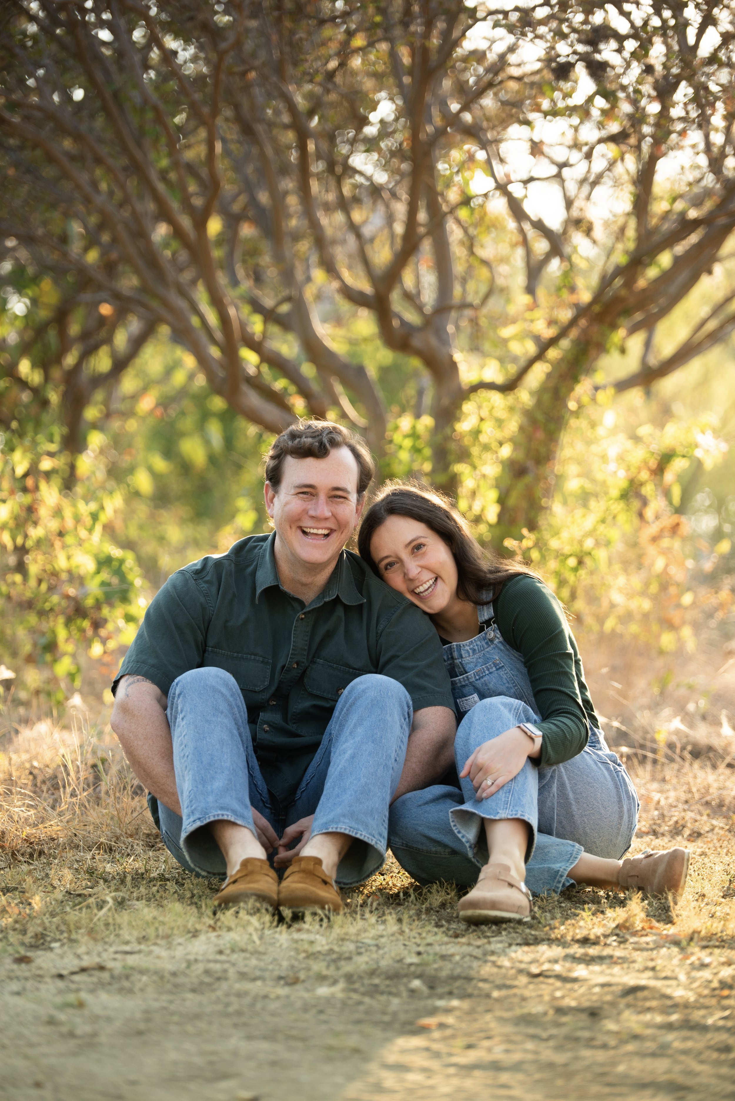 A smiling man and woman sitting on the ground outdoors, surrounded by trees with sunlight filtering through the leaves.