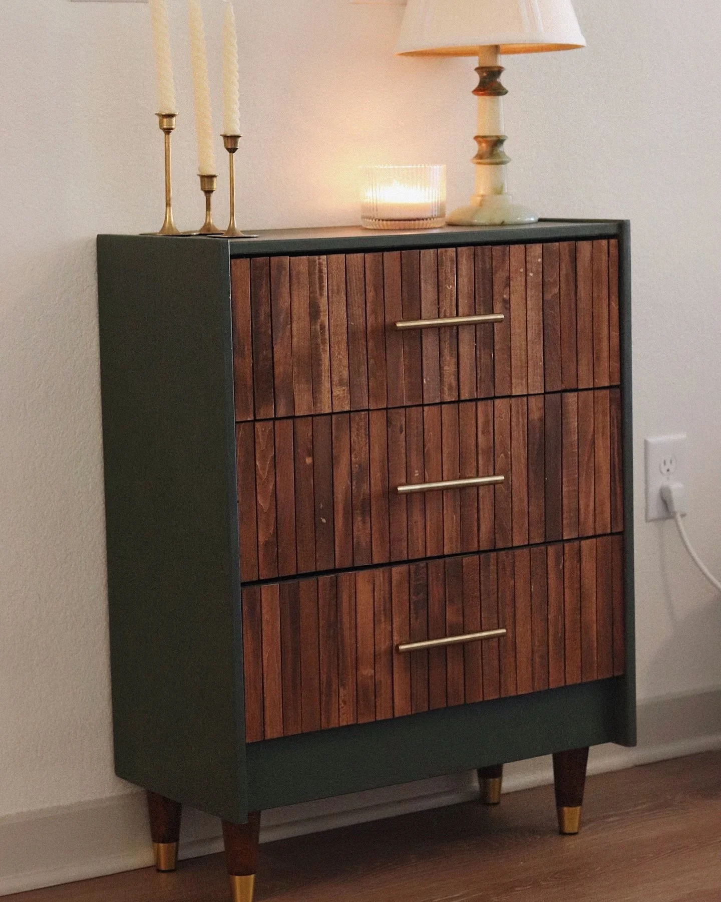 A vintage-style wooden dresser with four vertical drawers, a dark green frame, and angled legs with gold tips. On top, there are three gold candlesticks with white candles, a glass candle holder with a lit candle, and a cream-colored table lamp with 