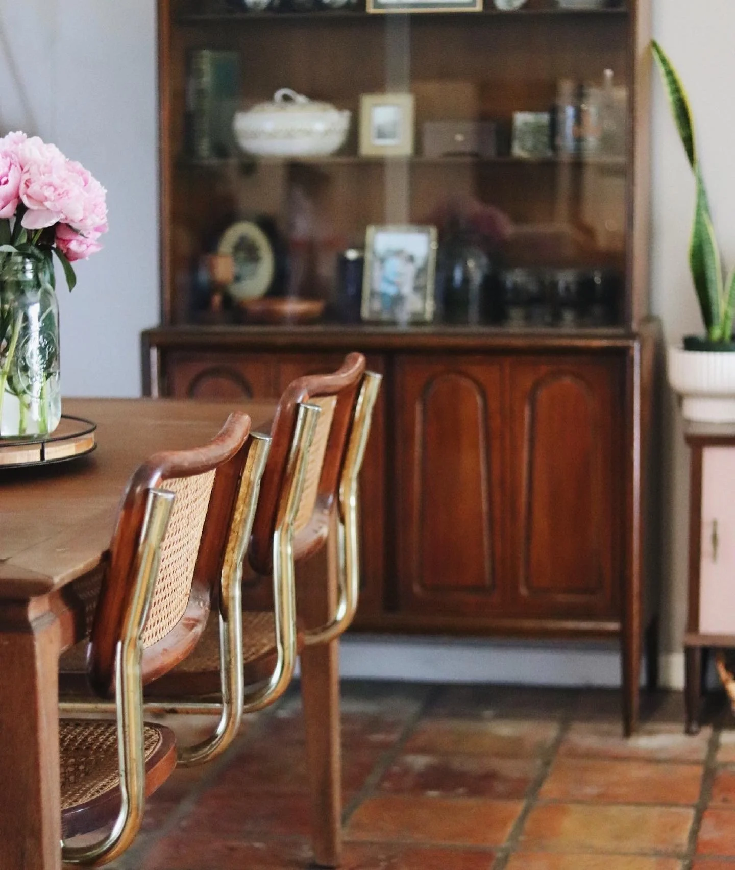A wooden dining table with a glass vase of pink peony flowers, surrounded by wooden chairs with woven seats, in front of a vintage wooden cabinet displaying decorative items and framed photographs.
