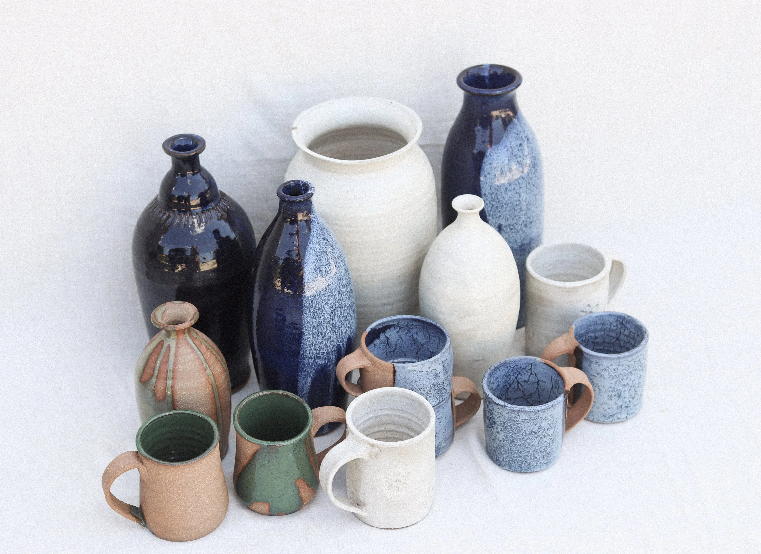 Collection of various ceramic vases and mugs in different shapes, sizes, and colors, some glazed and some unglazed, arranged on a white background.
