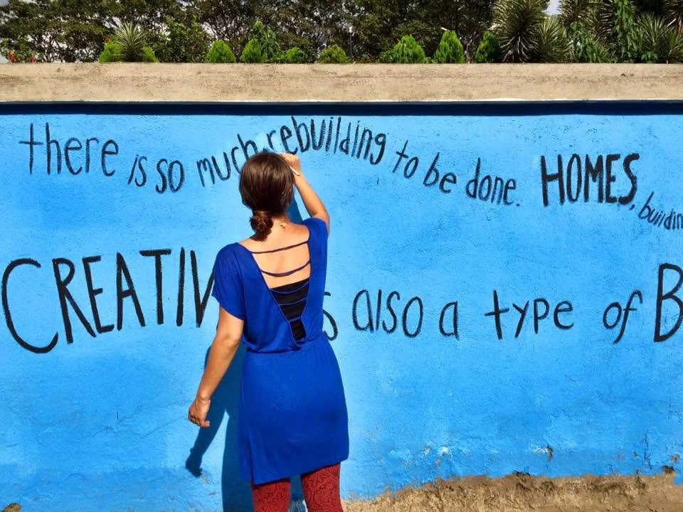 A woman writing on a blue wall with black text about rebuilding and creating, with trees and greenery above the wall.