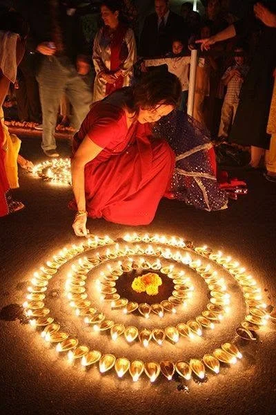 A woman in a red saree is participating in a traditional Nepali Diwali celebration, lighting diyas (oil lamps) arranged in concentric circles on the ground.