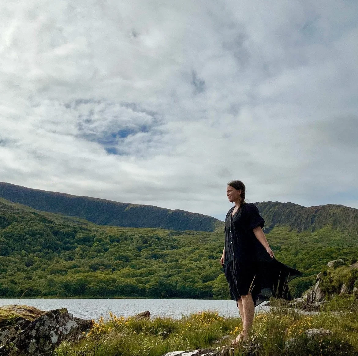 A woman standing on rocks near a lake with green hills and mountains in the background, and cloudy sky overhead.