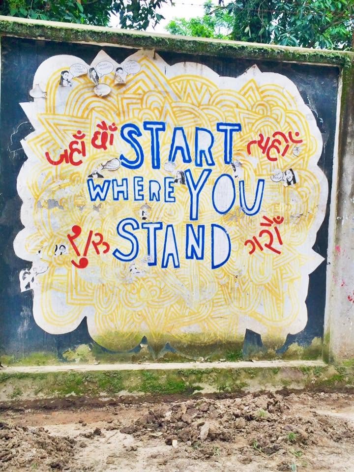Colorful mural with the quote "Start where you stand" in blue letters, surrounded by red and black Gujarati writing, on a wall with a yellow decorative background and some graffiti damage.
