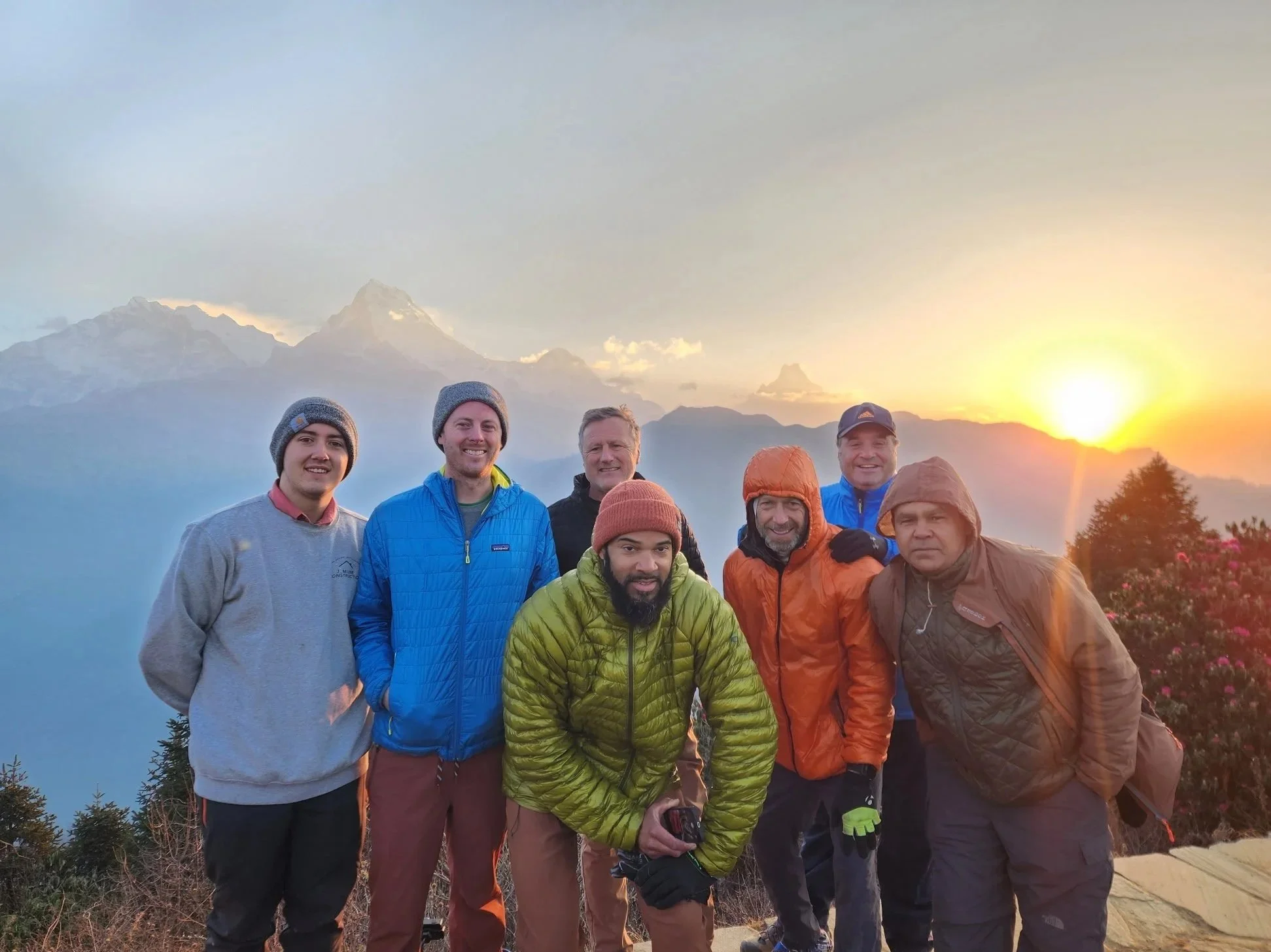 Group of seven men standing outdoors during sunset with mountains in the background, dressed in outdoor and hiking gear including jackets, hats, and beanies.