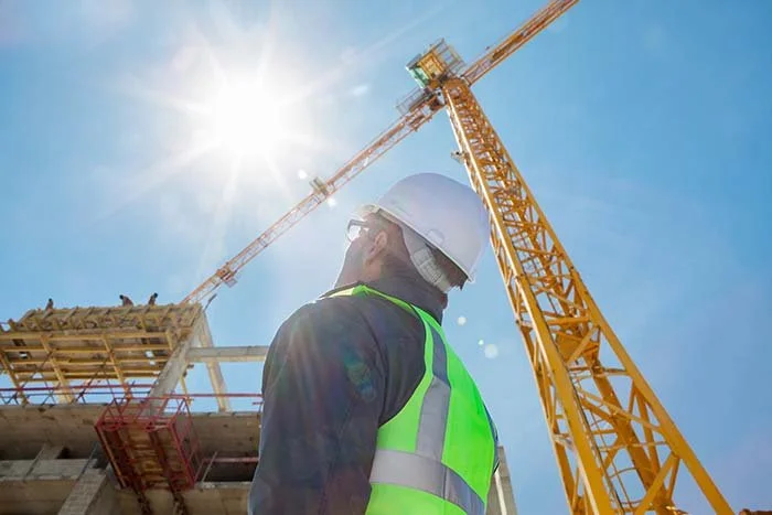 Construction worker wearing a helmet and neon safety vest stands at a construction site with a large yellow tower crane, under a bright sunny sky.