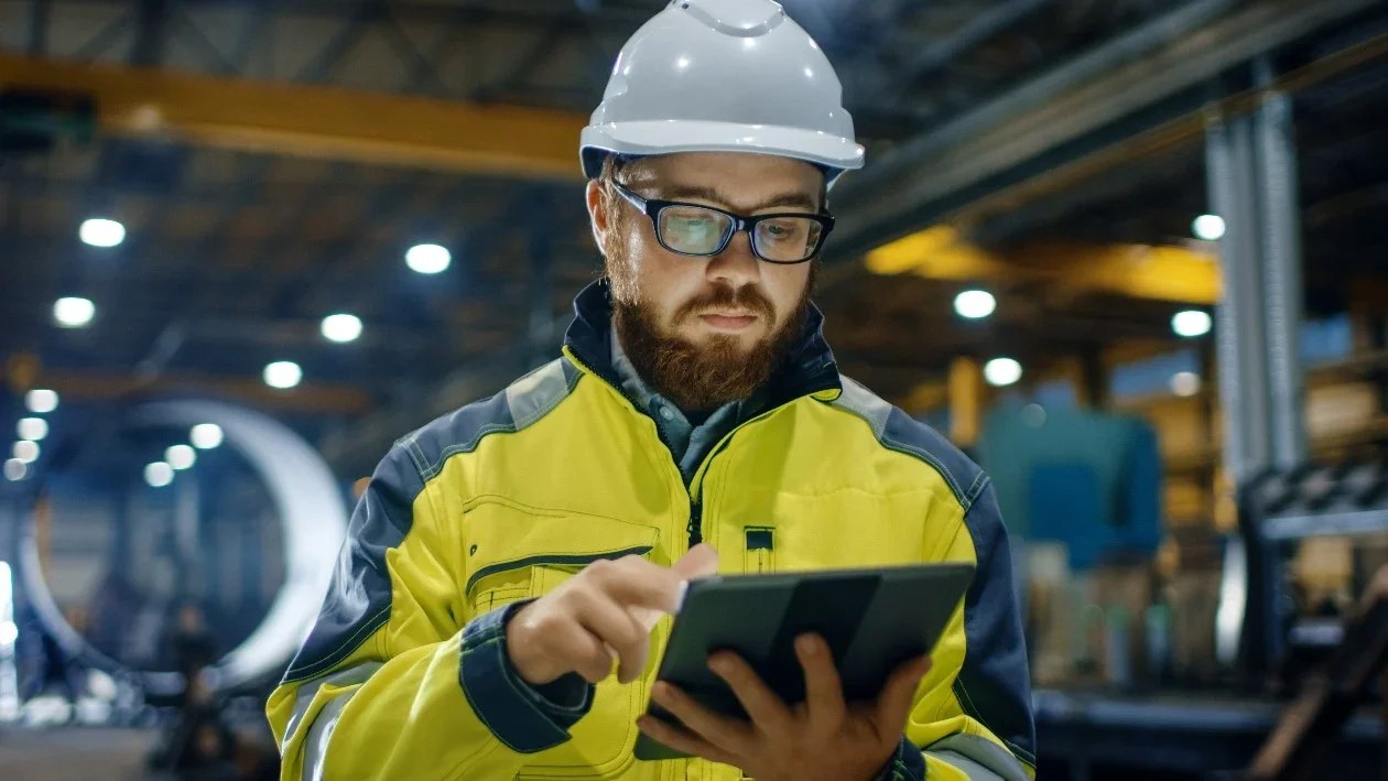 Man in safety gear inspecting equipment in an industrial warehouse using a tablet.
