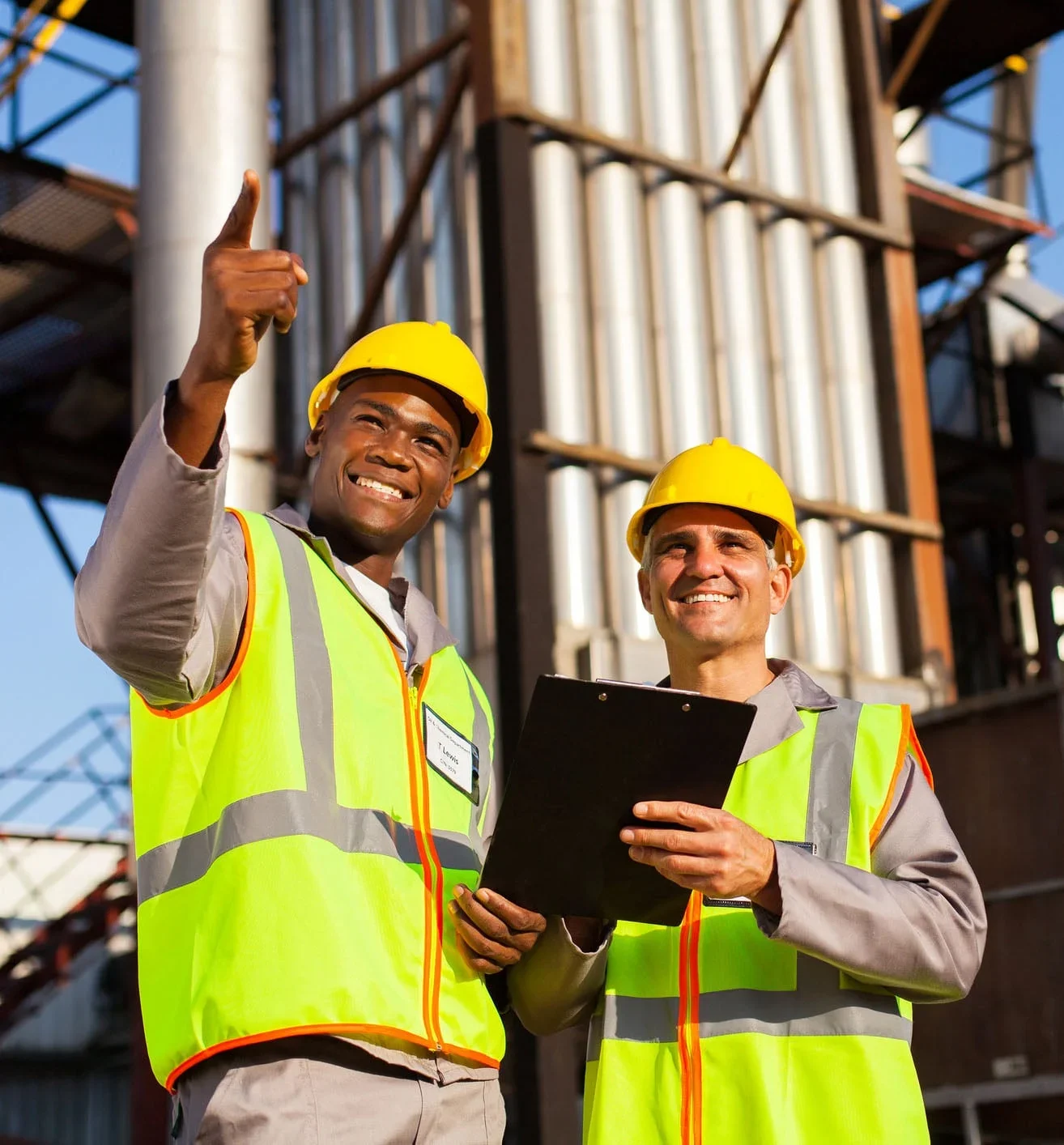 Two construction workers wearing yellow safety helmets and high-visibility vests smiling at the construction site, with one pointing upwards and the other holding a clipboard.